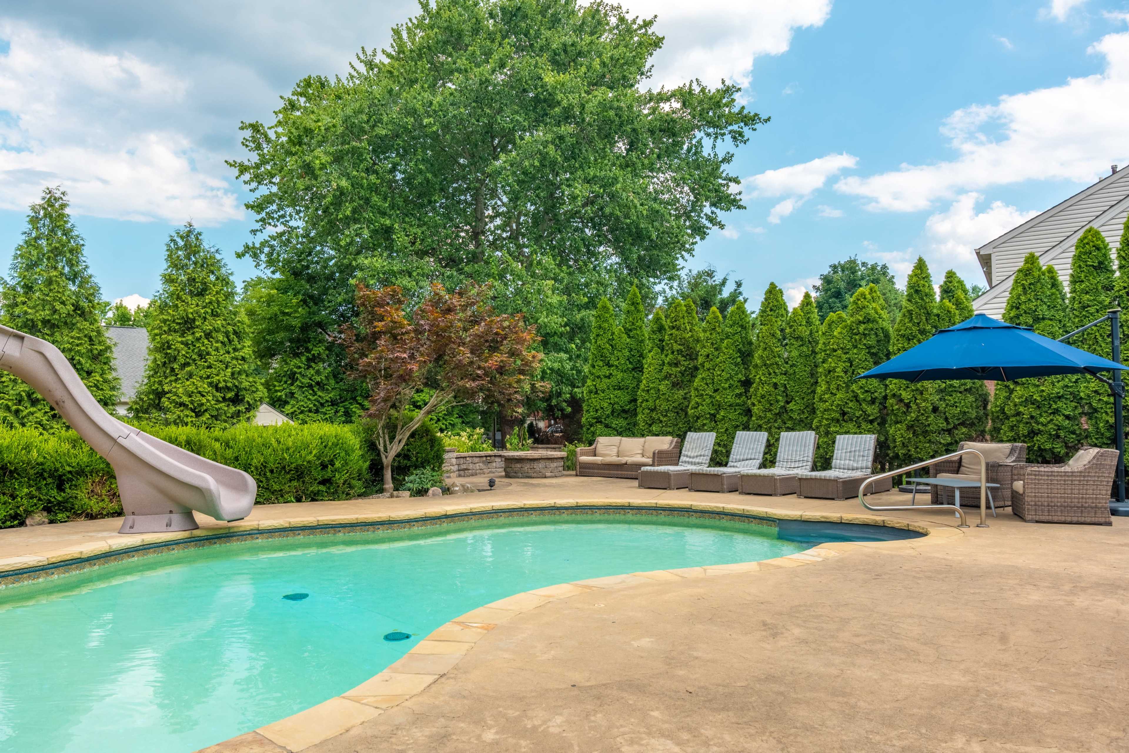 A backyard with a swimming pool, a water slide, lounge chairs, and a shaded seating area surrounded by tall green trees.
