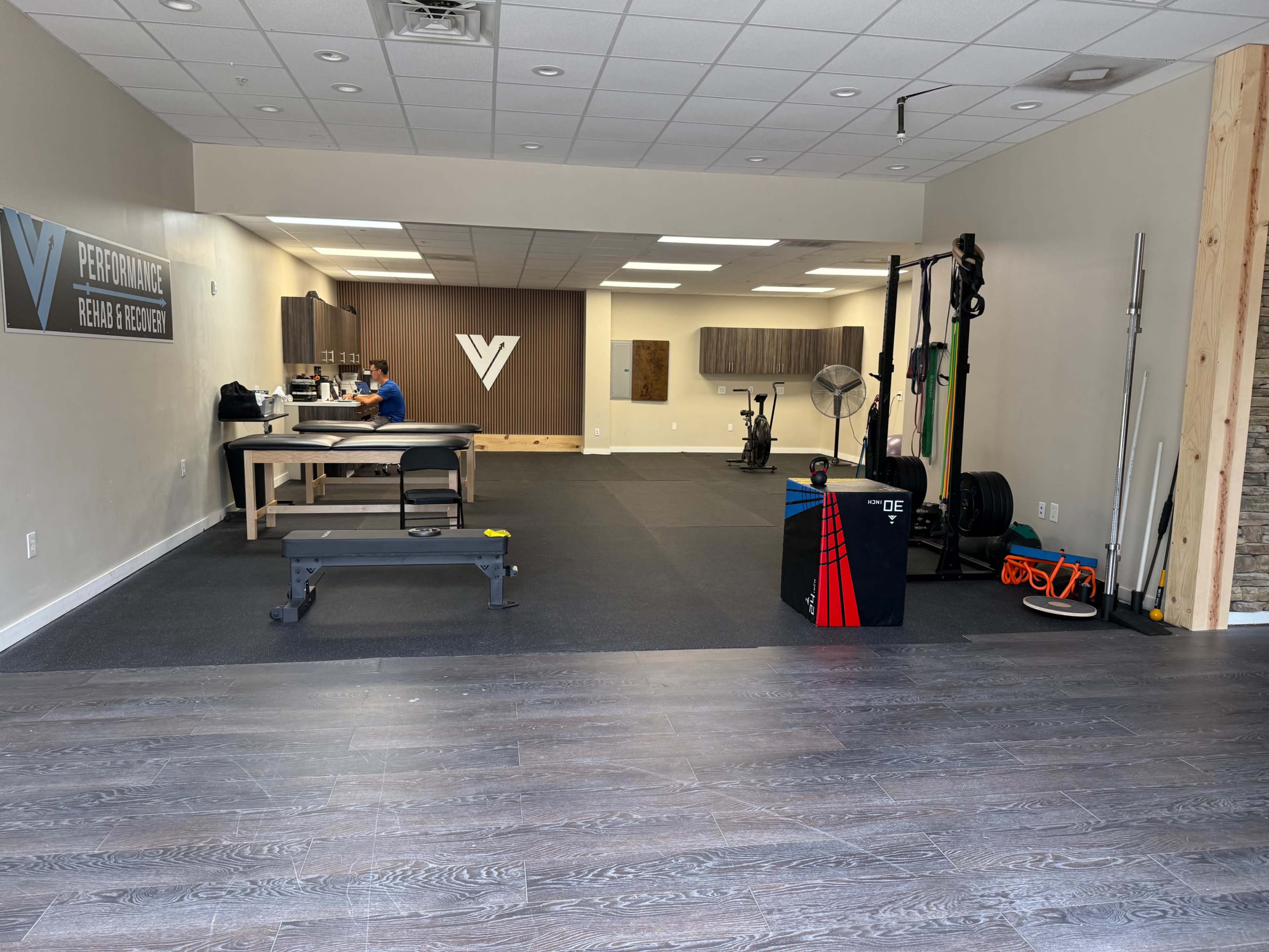 The image shows a fitness and rehabilitation space with workout equipment, a treatment table, and a person seated at a desk.