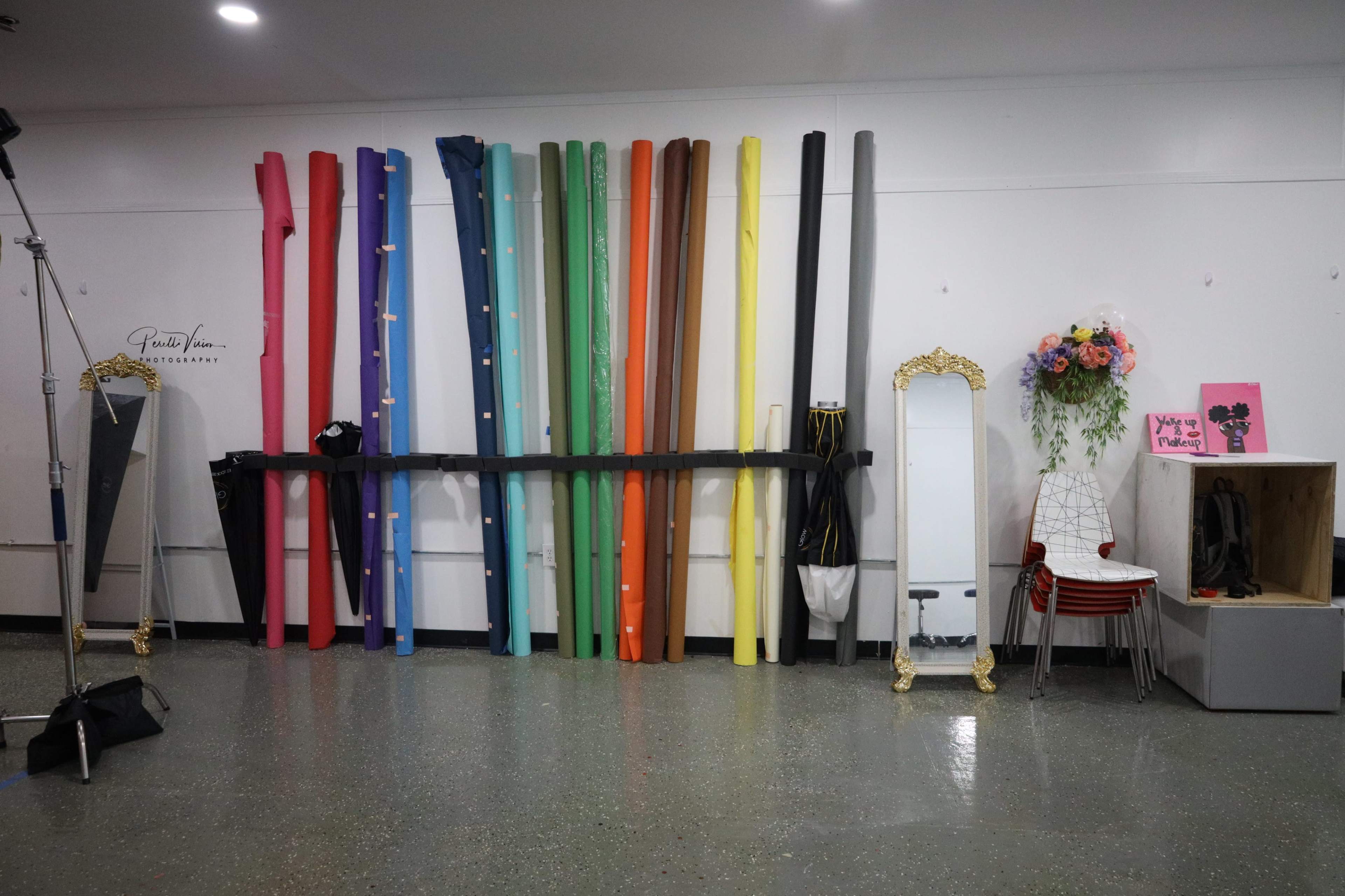 The image shows a studio space with colorful backdrop rolls on a wall, a full-length mirror, and a bouquet of flowers displayed nearby.