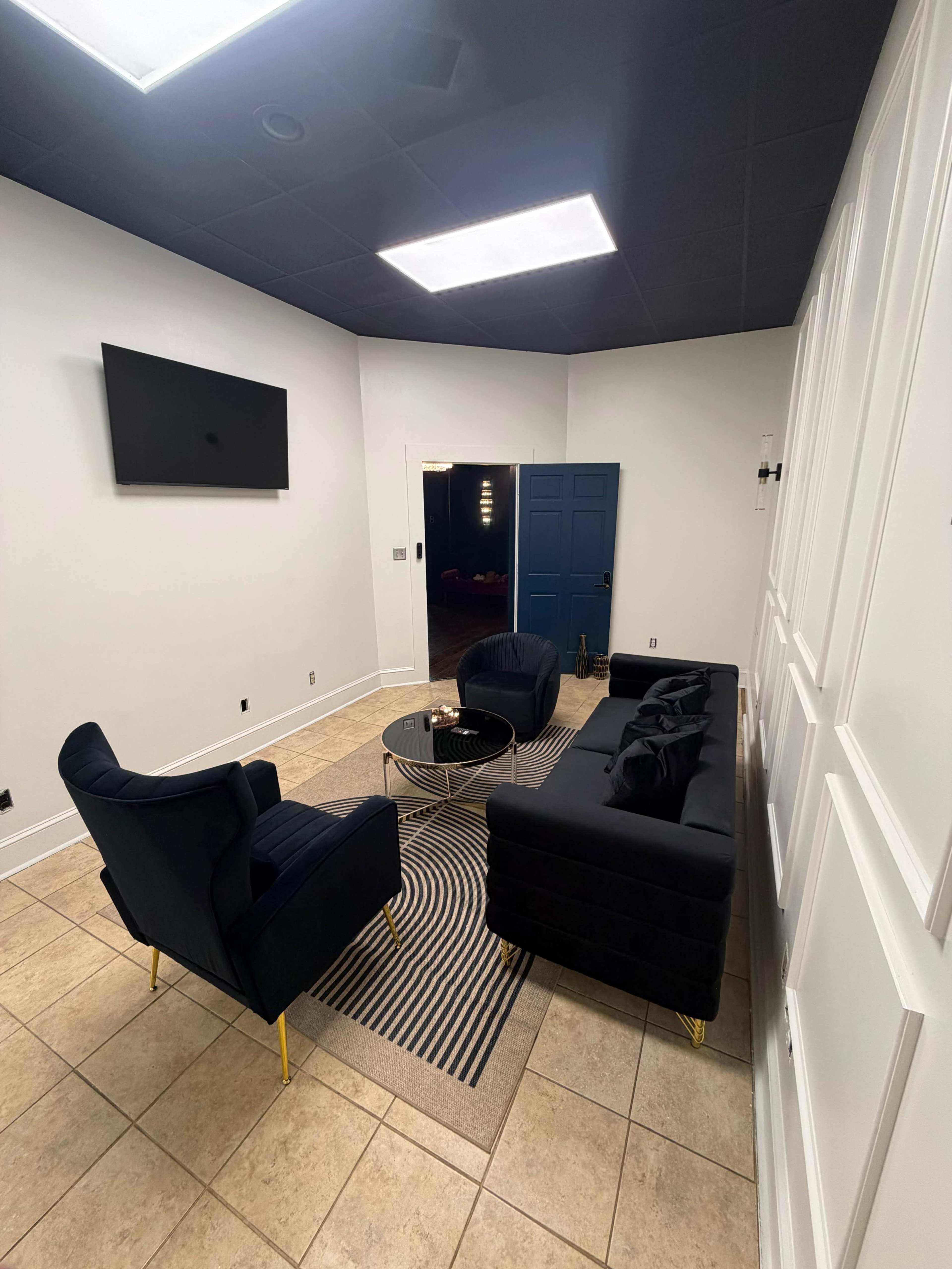 The image shows a small room with two black sofas facing each other over a round coffee table, a television mounted on the wall, and a blue door leading to another space.