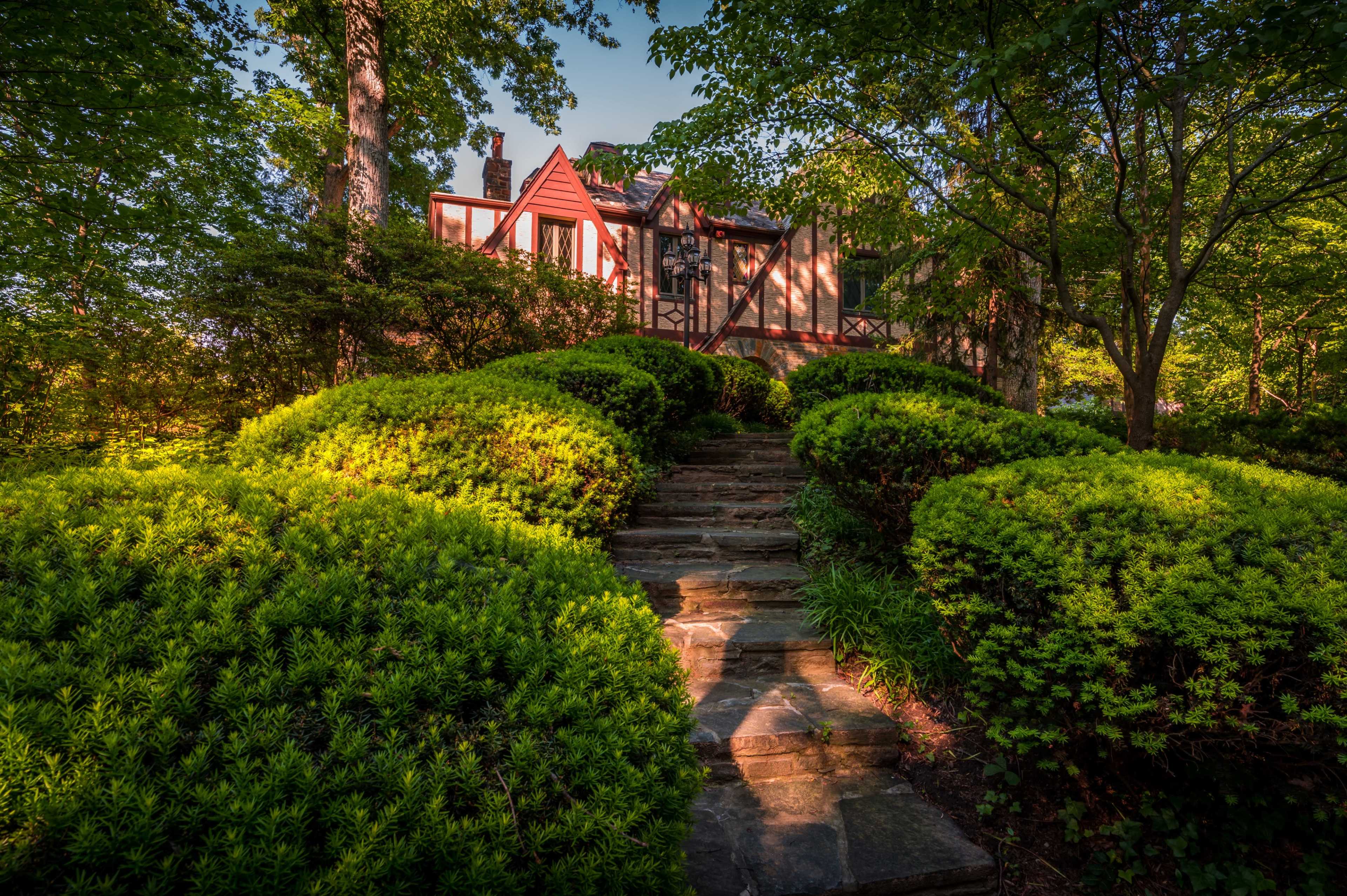 A stone pathway leads through lush green shrubs to a house partially obscured by trees.