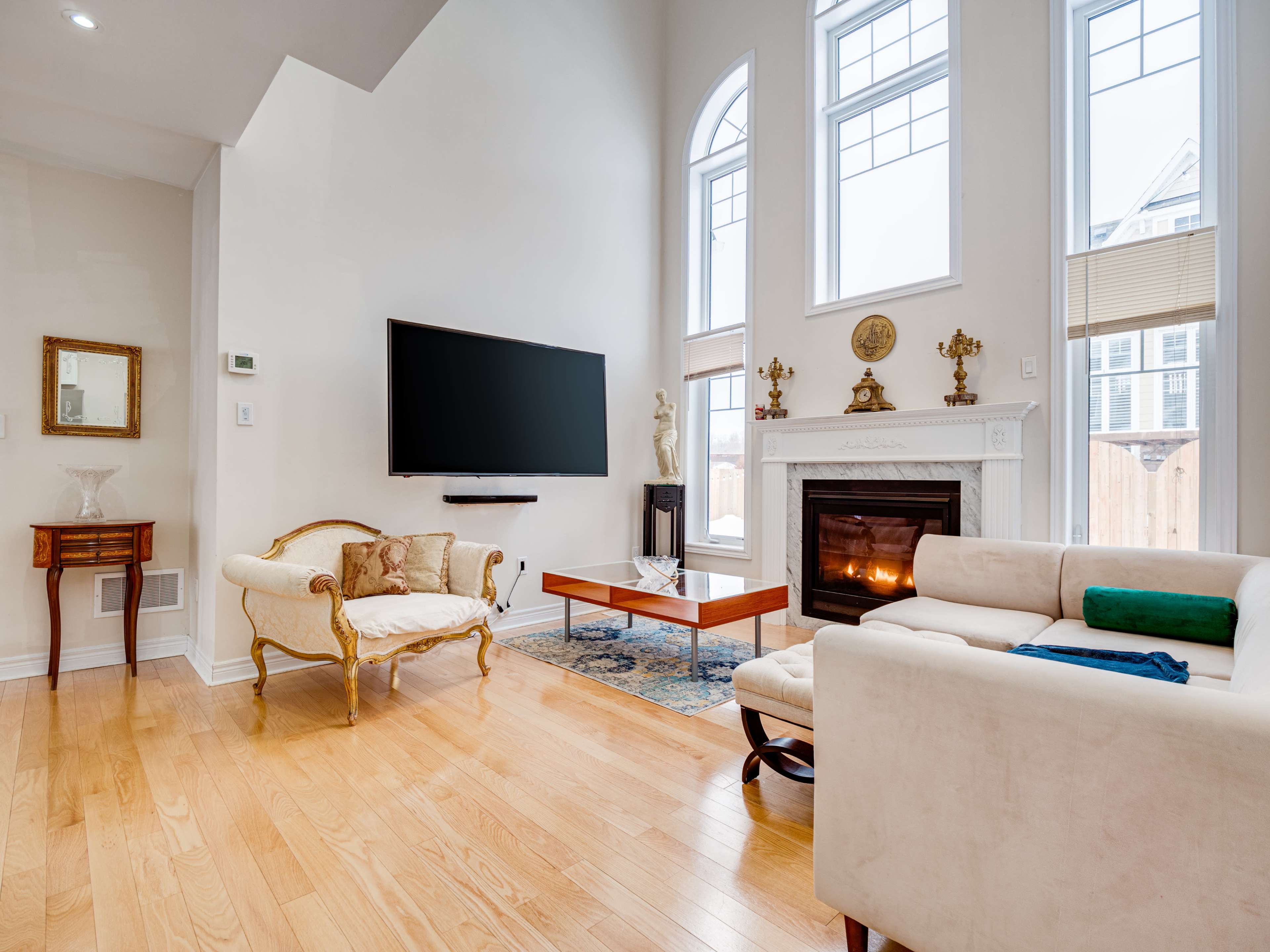 The scene features a living room with hardwood floors, a modern television mounted on the wall, a fireplace with a mantel, and a mix of contemporary and traditional furniture.