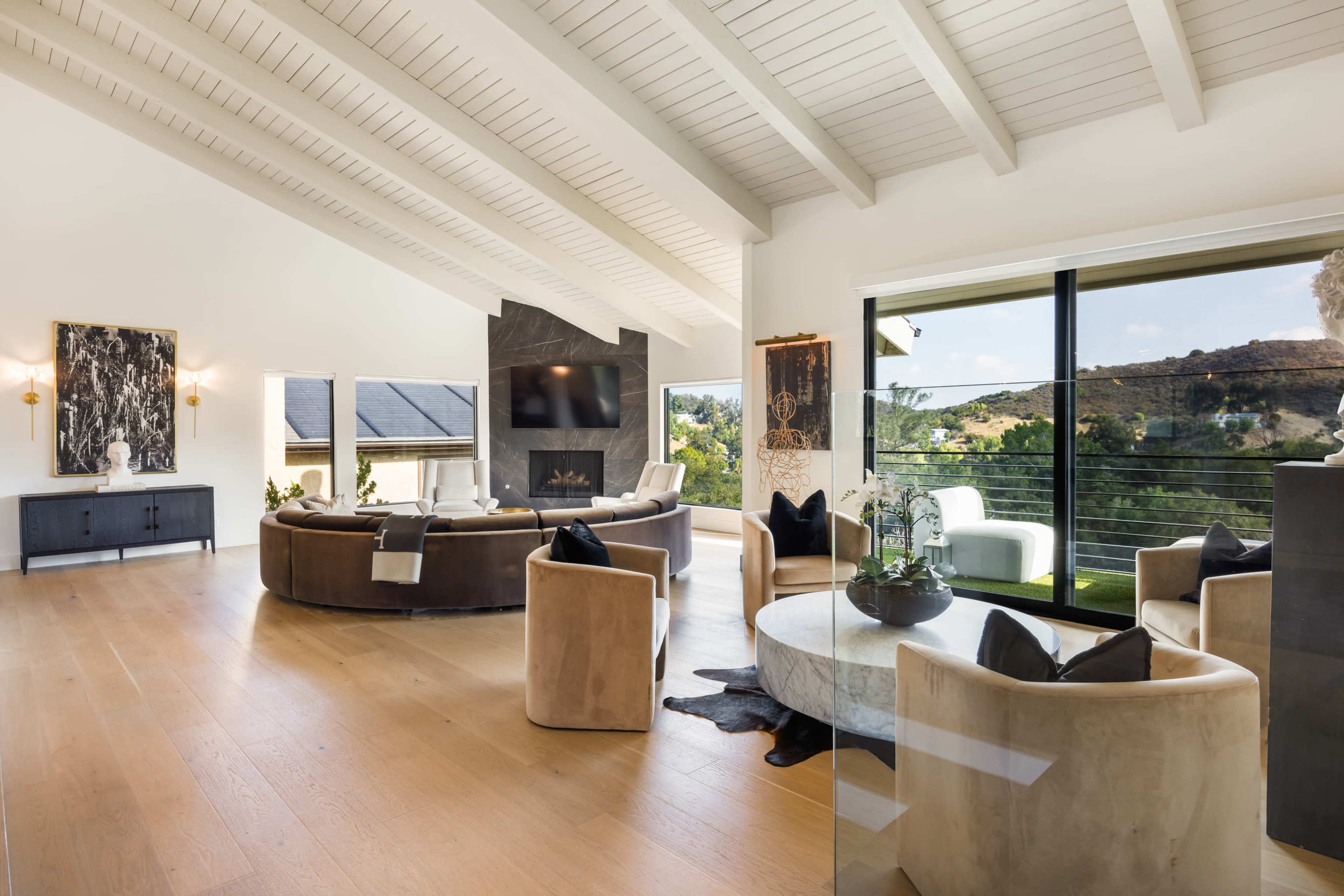 The image shows a modern living room with high, sloped ceilings, large windows overlooking a landscape, and circular seating arranged around a central coffee table.
