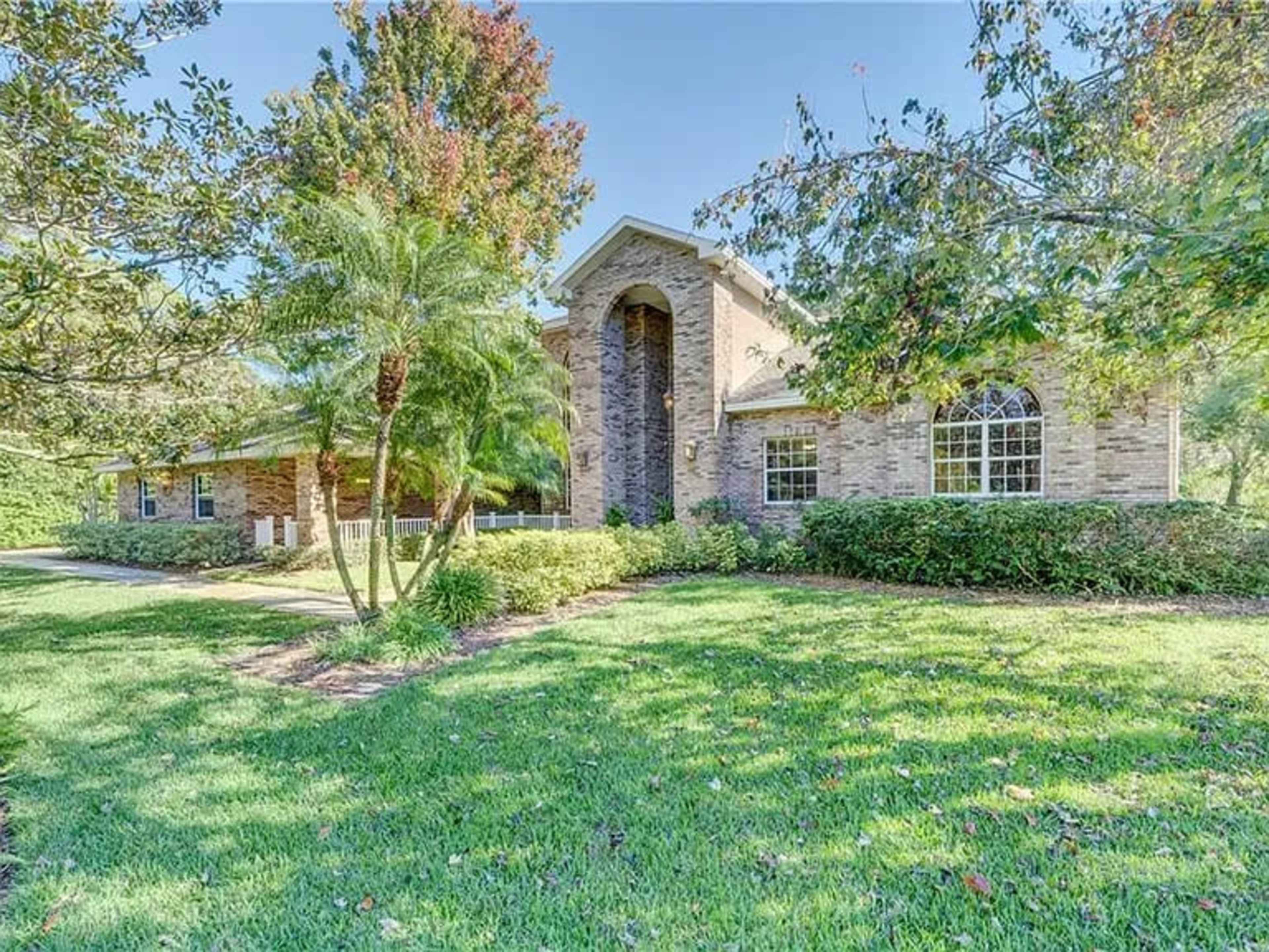 A brick house with a large front porch and tall entrance is surrounded by green lawn and trees.