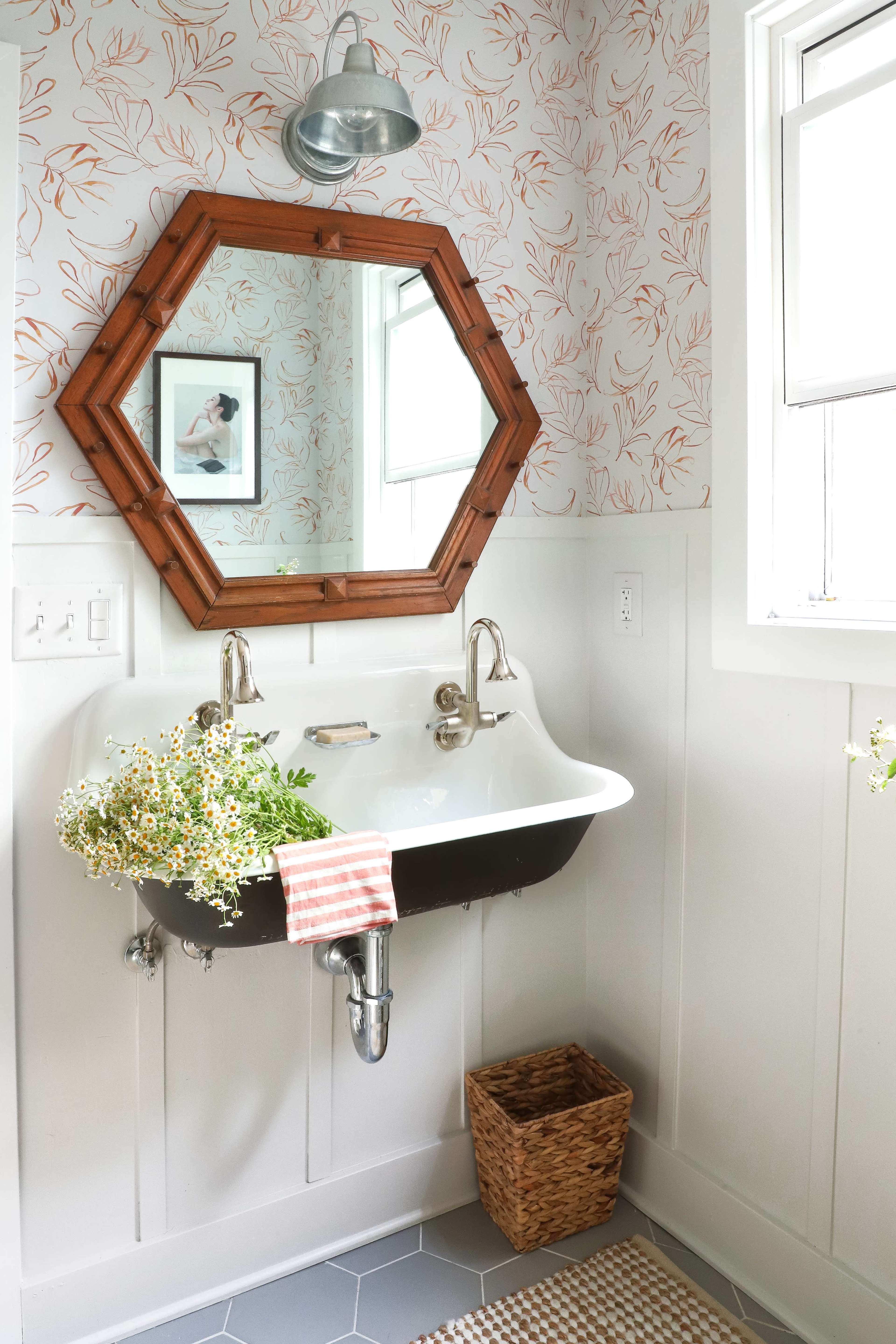 A small bathroom with an octagonal wooden mirror above a wall-mounted sink, surrounded by floral wallpaper and a woven basket on the floor.