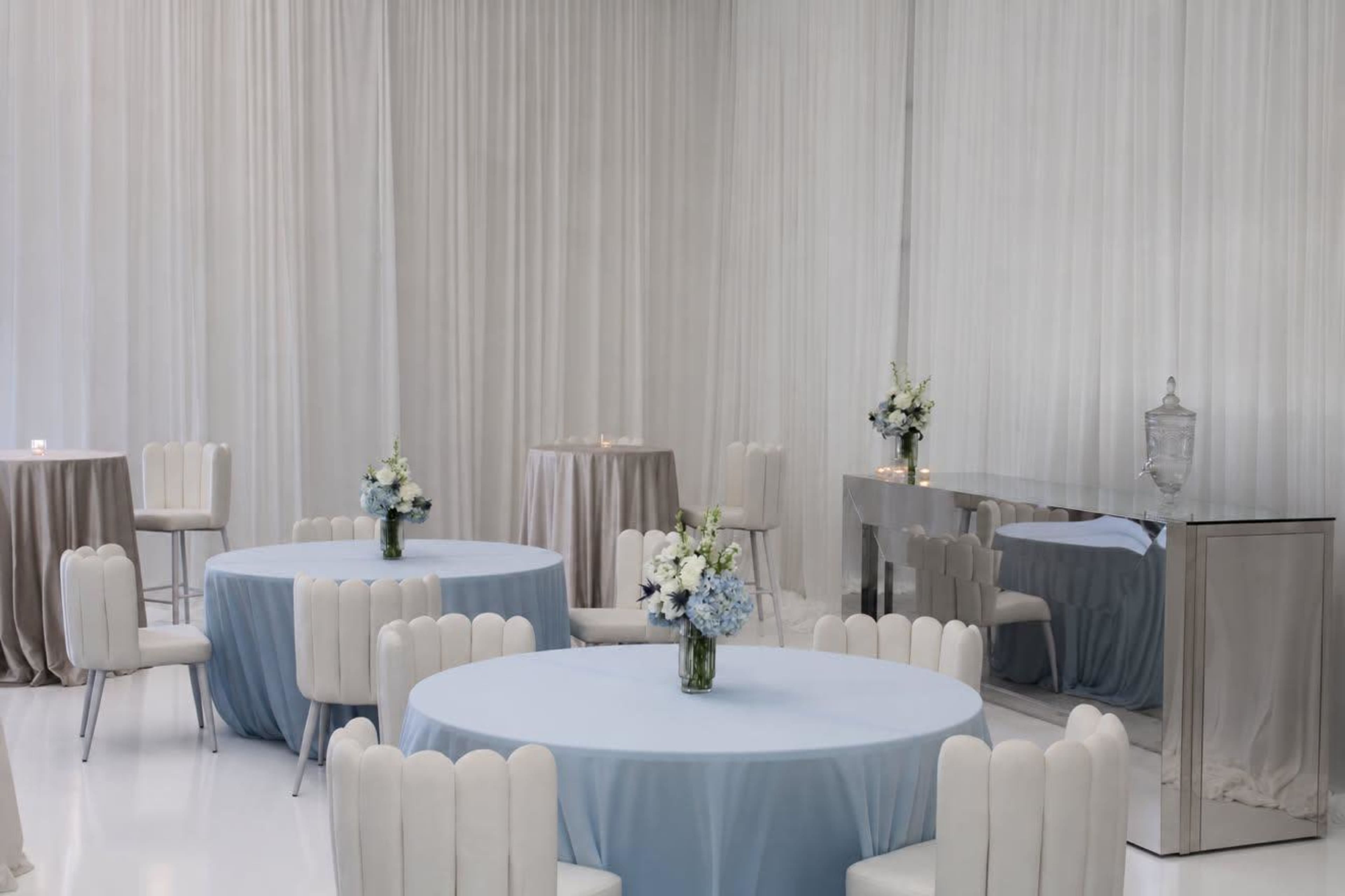 The scene features a bright, modern event space with several round tables covered in light blue tablecloths, surrounded by white chairs, and adorned with floral centerpieces.