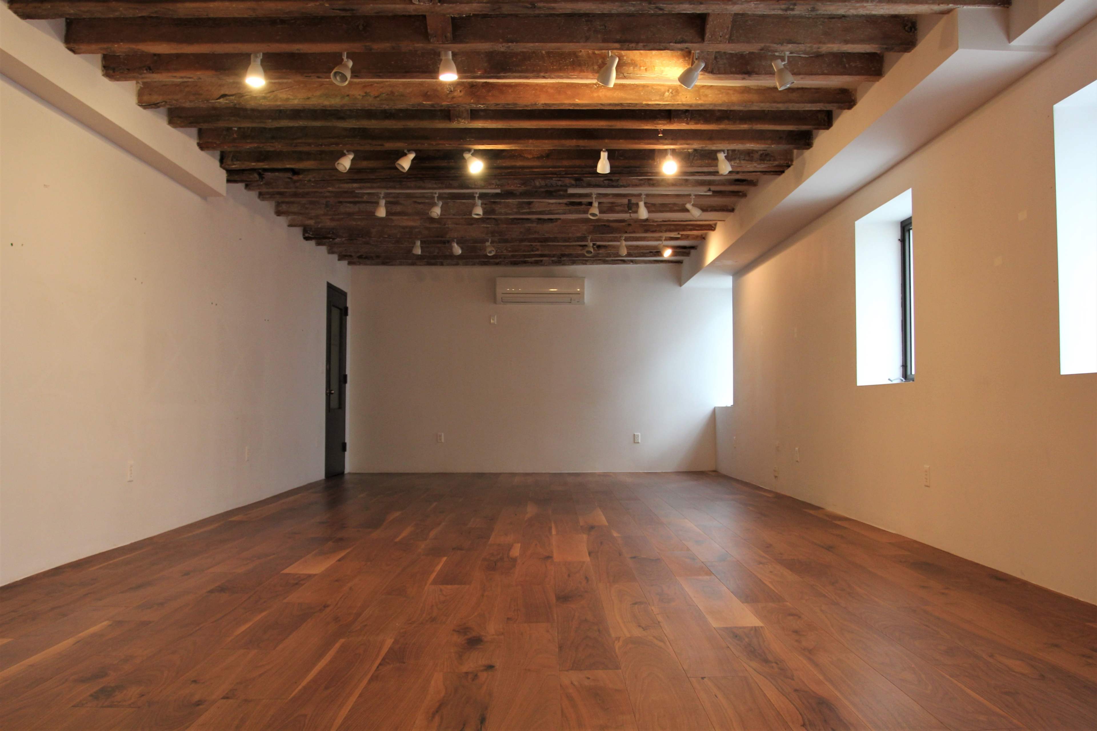An empty room features wooden flooring, exposed beam ceilings, and two windows allowing natural light.