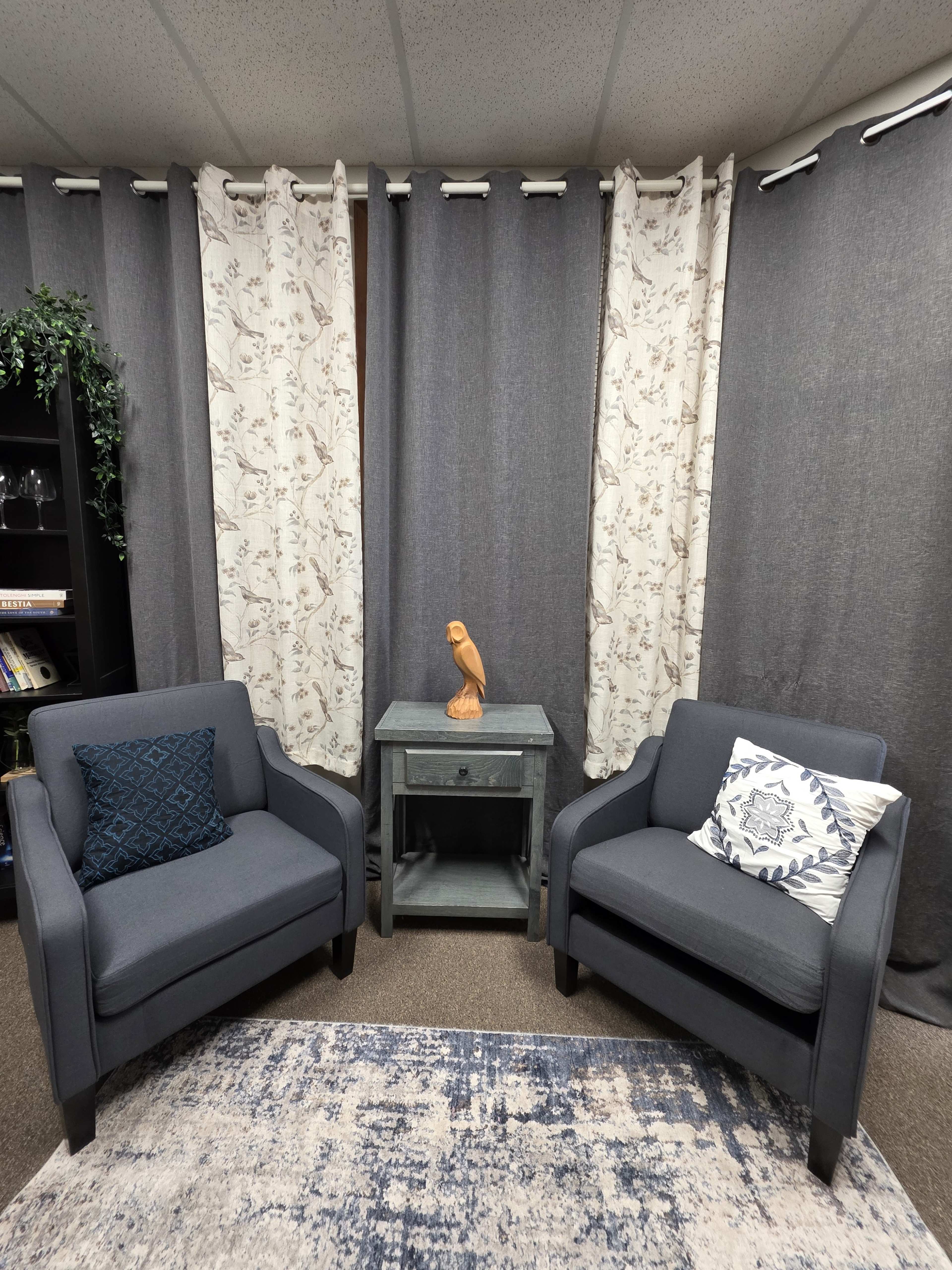 The image shows two gray armchairs positioned around a small wooden side table, with patterned curtains and a decorative bird sculpture on the table.
