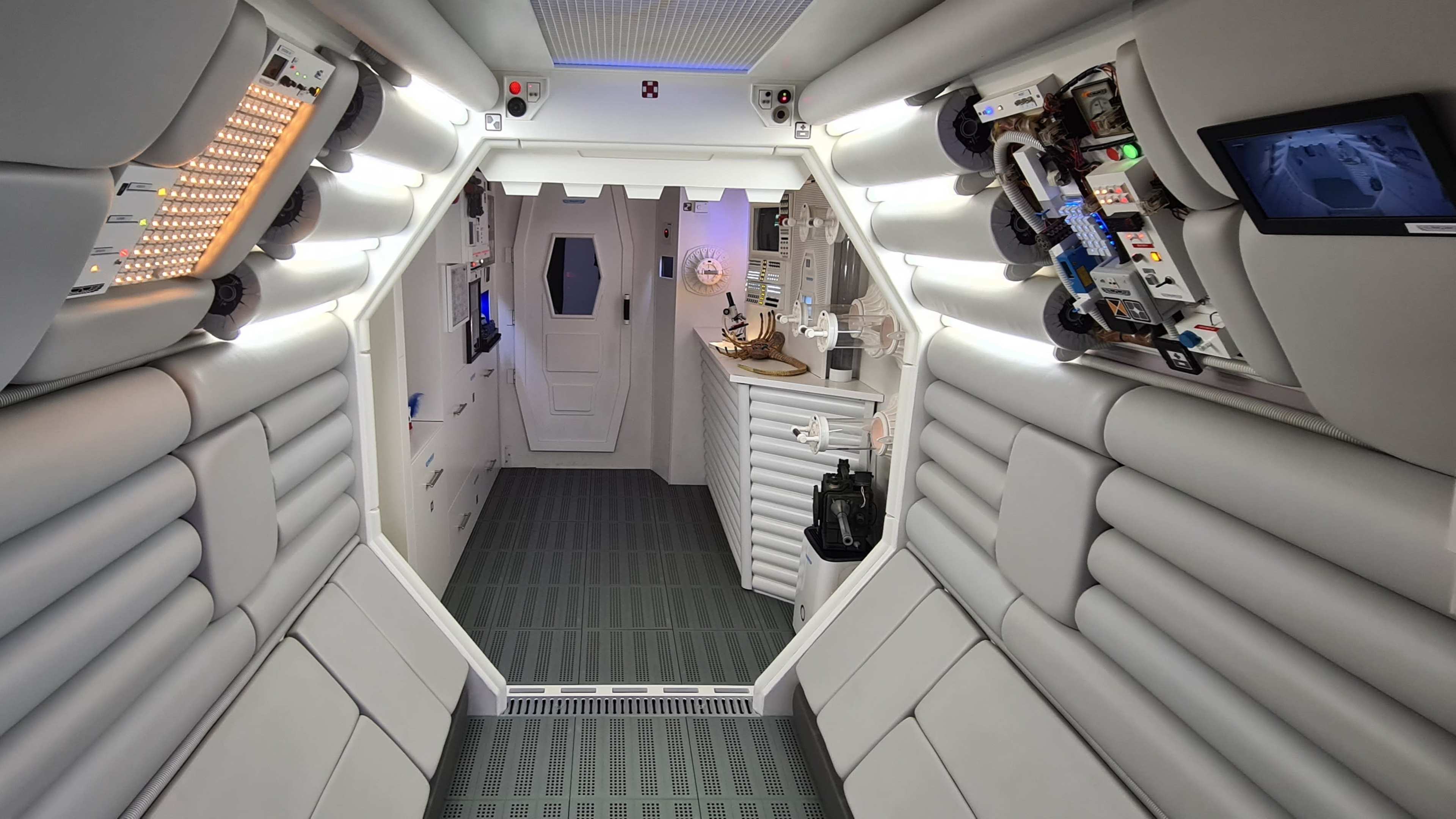The interior of a futuristic spacecraft corridor, featuring curved white walls, various control panels, and a central access door.