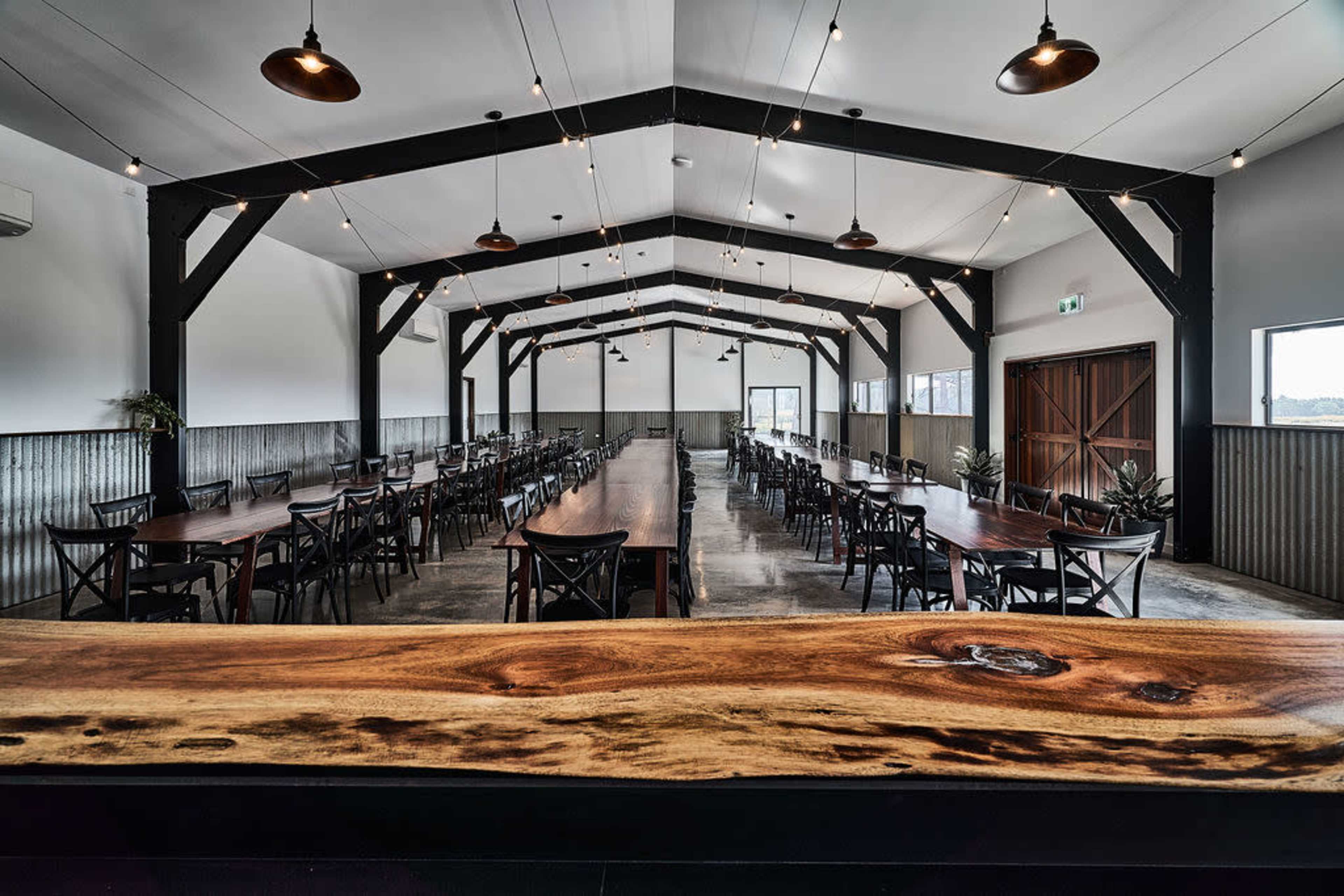A large dining hall with long tables arranged in rows under a high ceiling supported by wooden beams, illuminated by overhead lights.