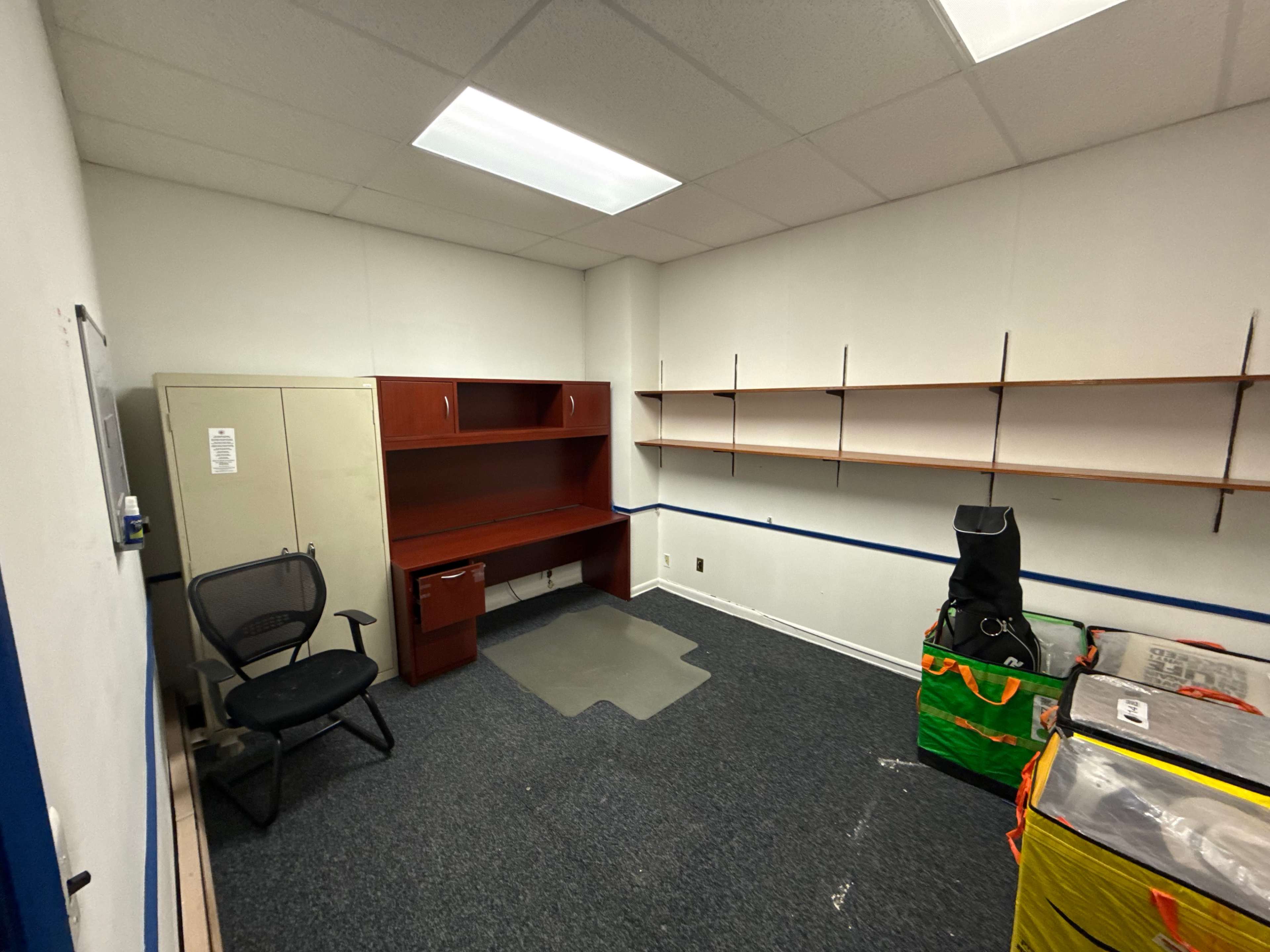 The image shows an empty office space featuring a desk, a chair, and shelving on the walls, with some storage bins in the corner.