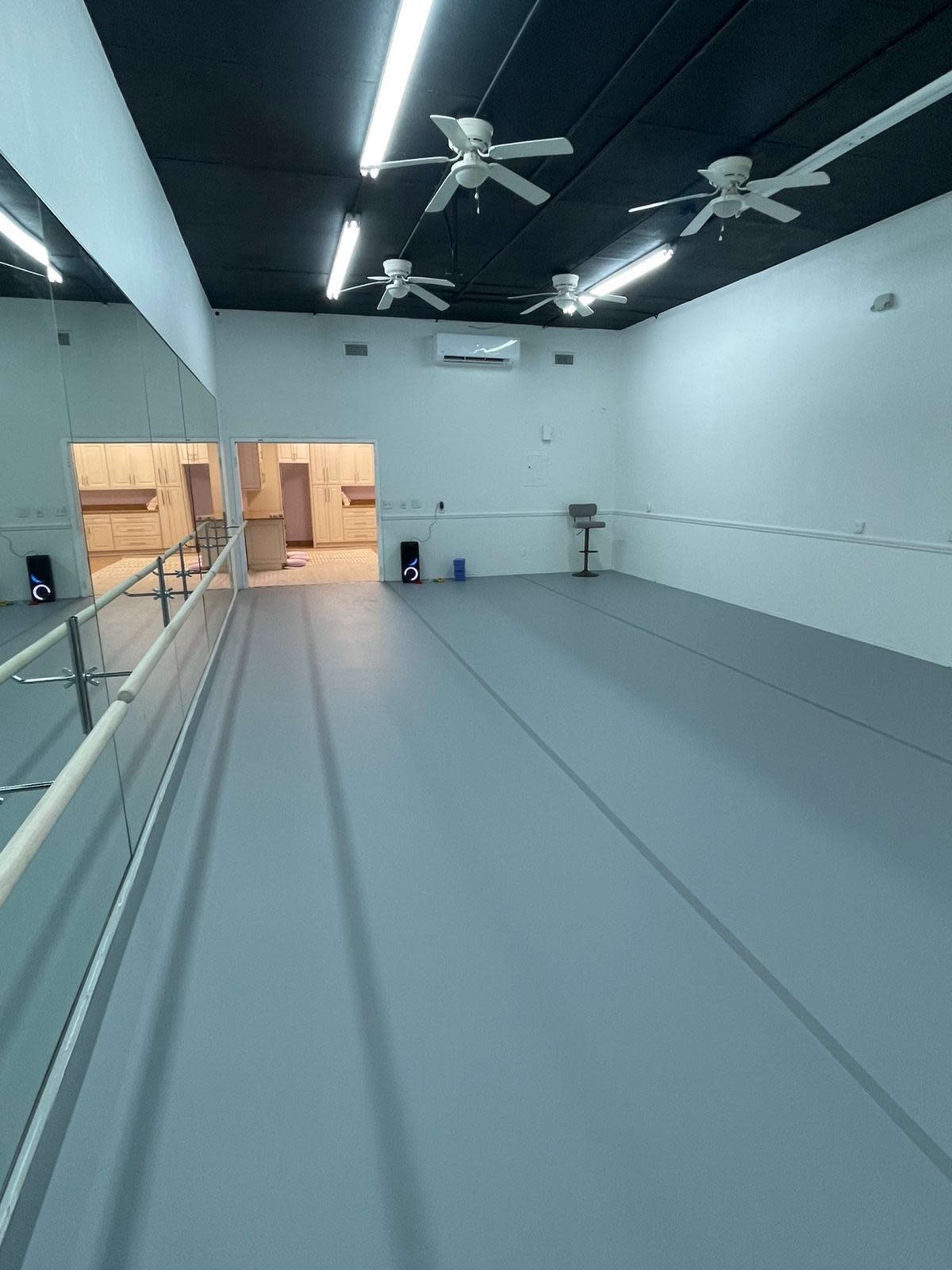 The image shows an empty, well-lit dance studio featuring a large mirror on one wall and gray flooring.