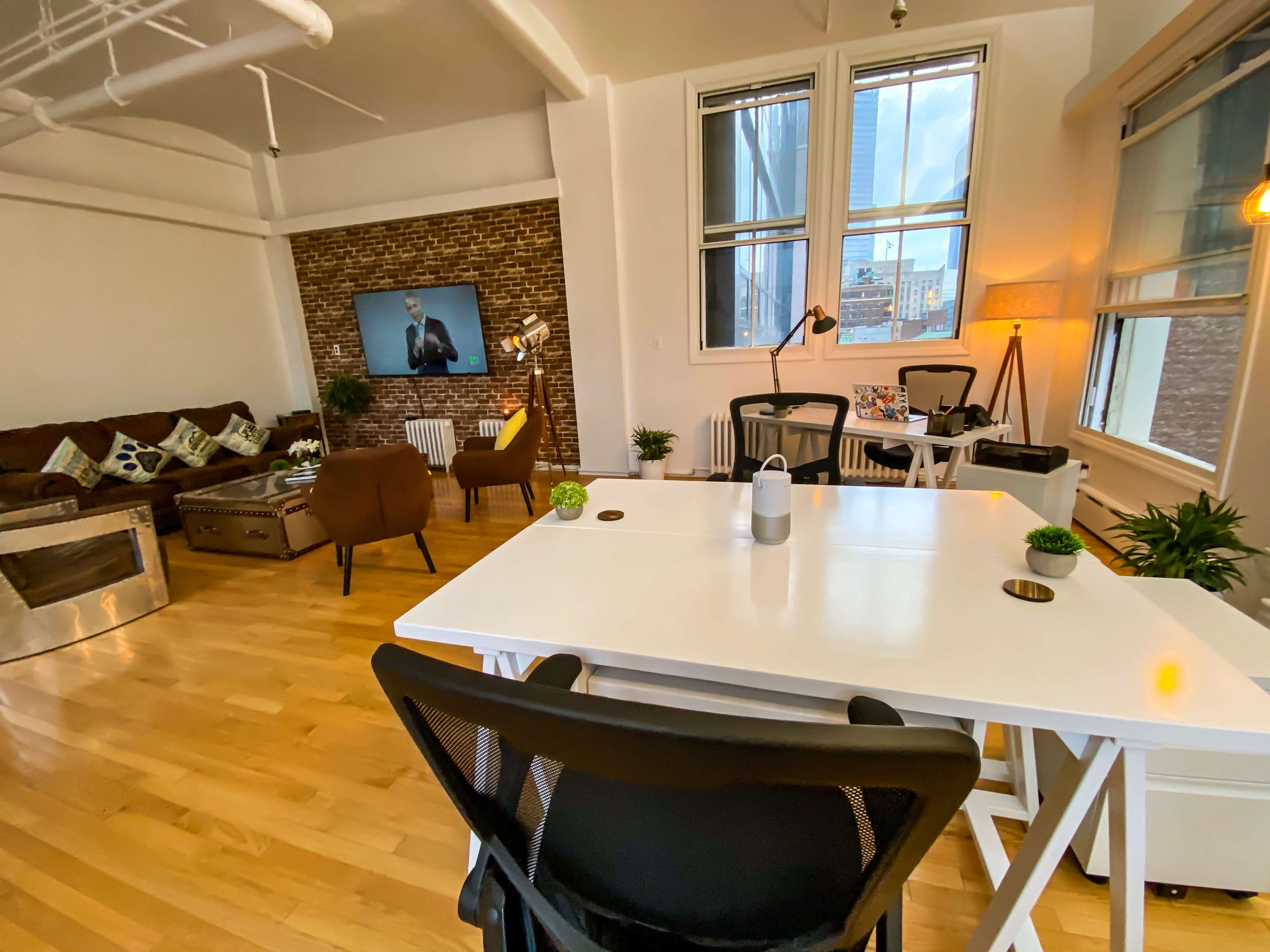 A modern open-concept living space with a desk area, a couch, and a television mounted on the brick wall.