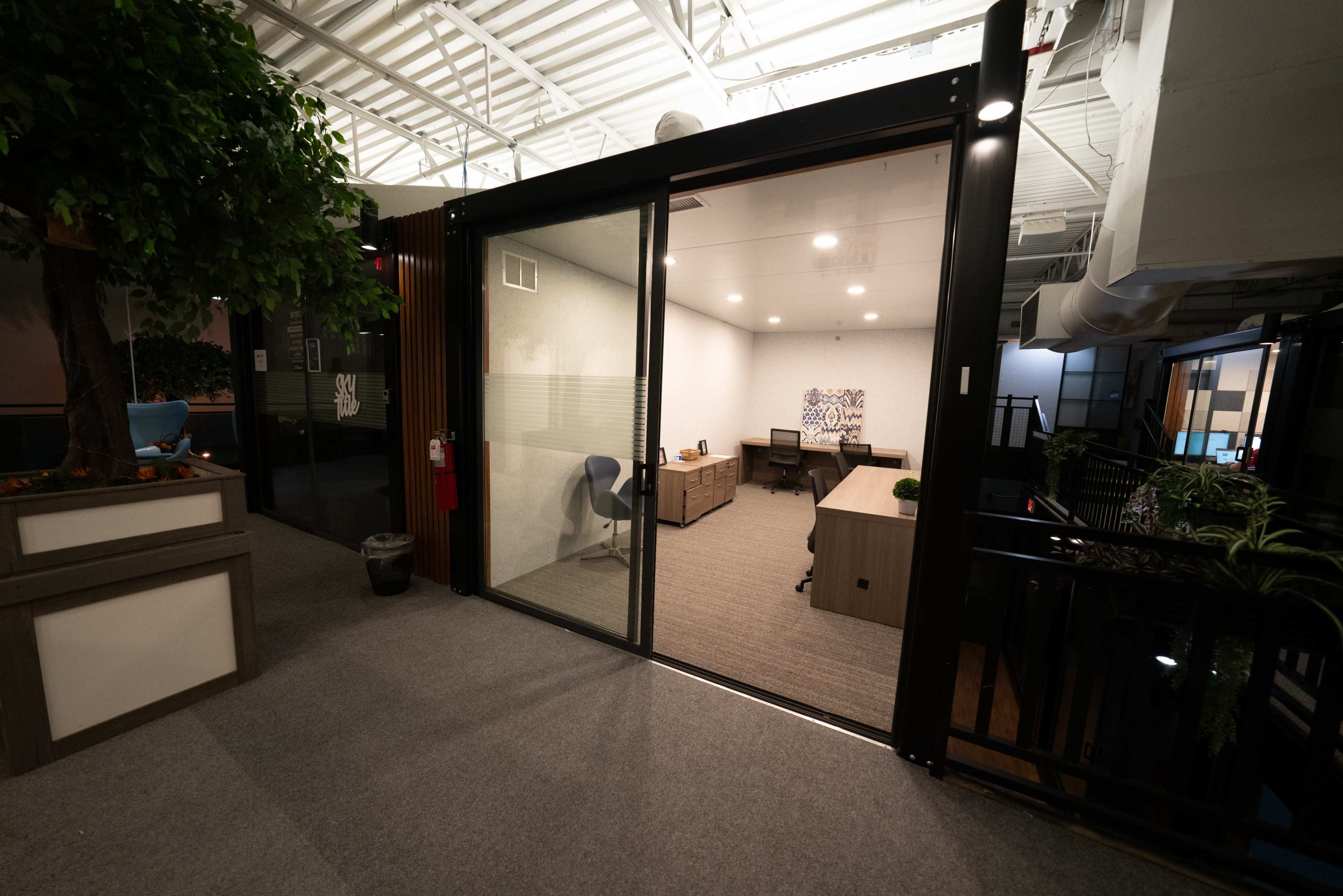 The image shows a modern office space with glass walls, featuring a desk, chair, and decorative plants inside.