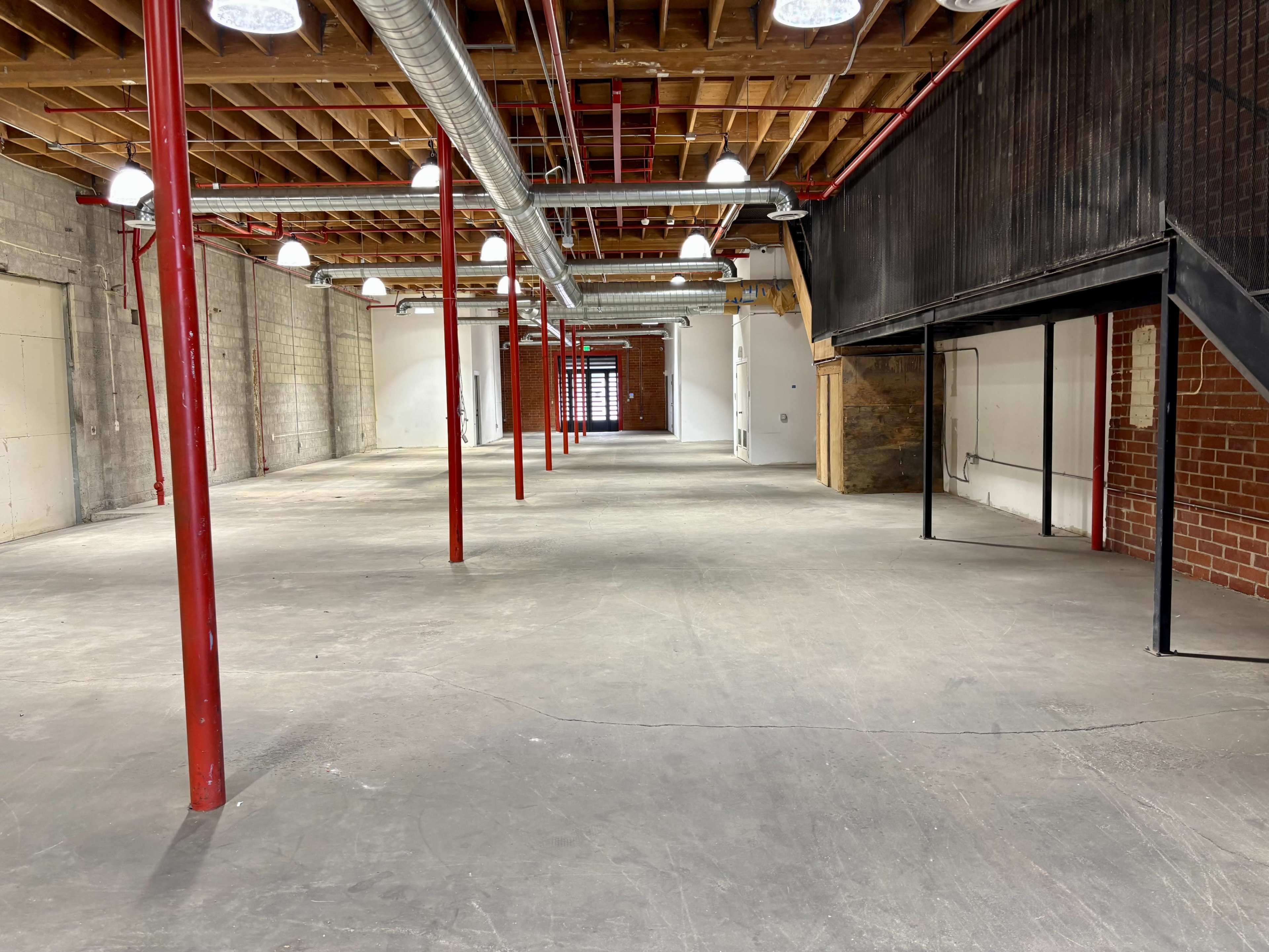 An empty industrial space features concrete floors, exposed bricks, and red metal support columns under a wooden ceiling.