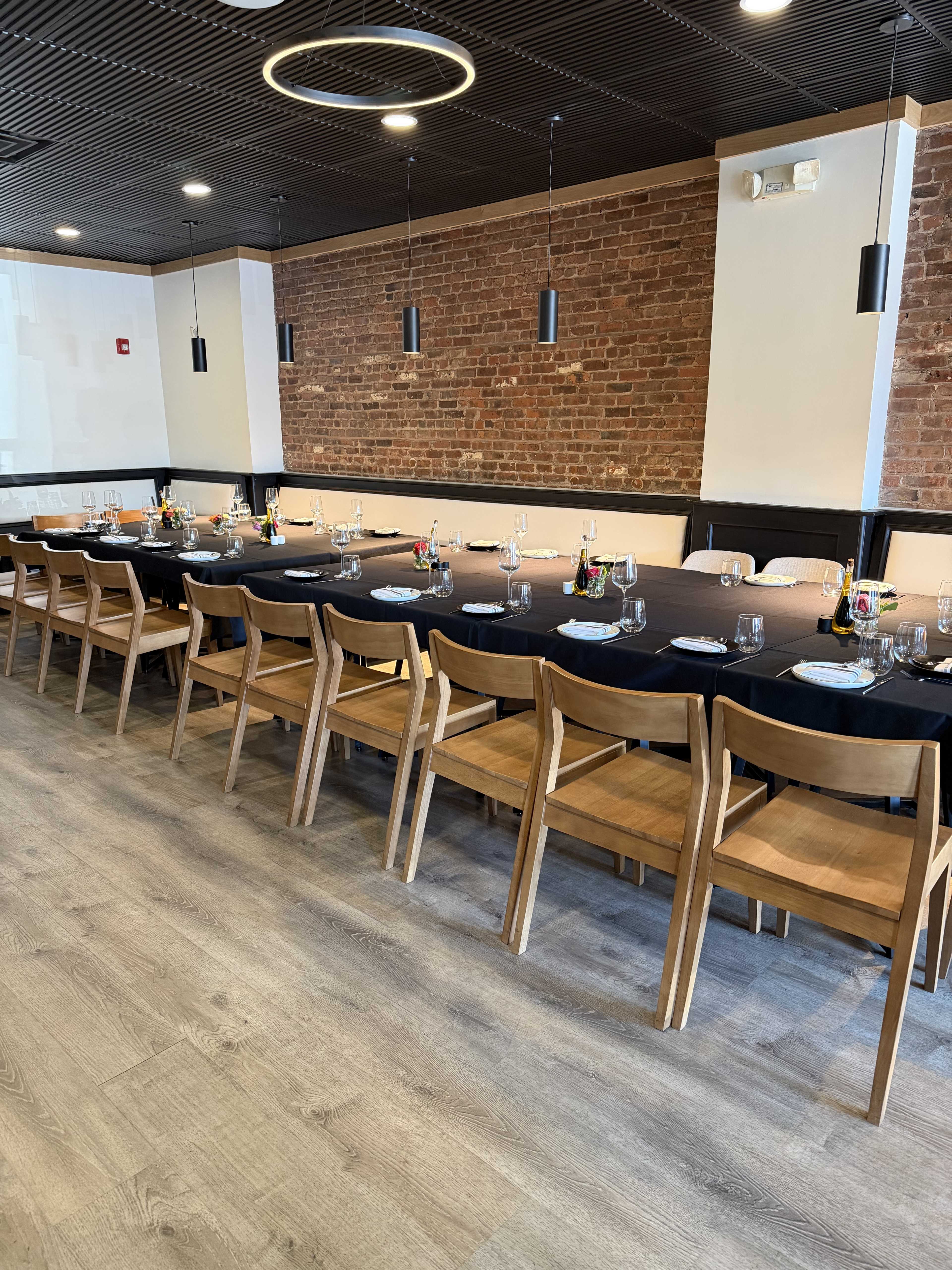 The image shows a dining area with a long table set for a meal, surrounded by wooden chairs and against a backdrop of exposed brick walls.