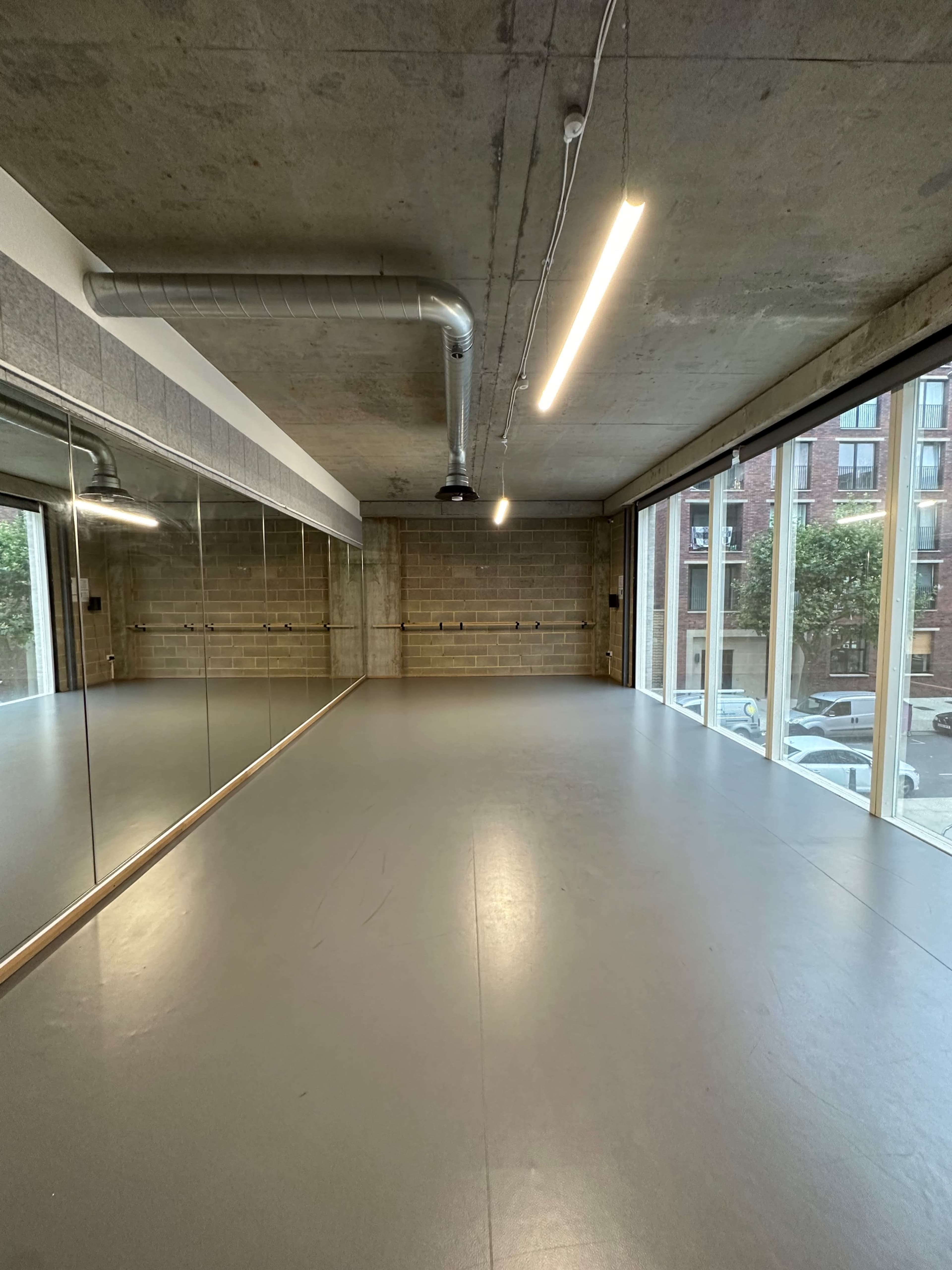 An empty dance studio with a large mirror, concrete walls, and large windows revealing a view of parked cars outside.