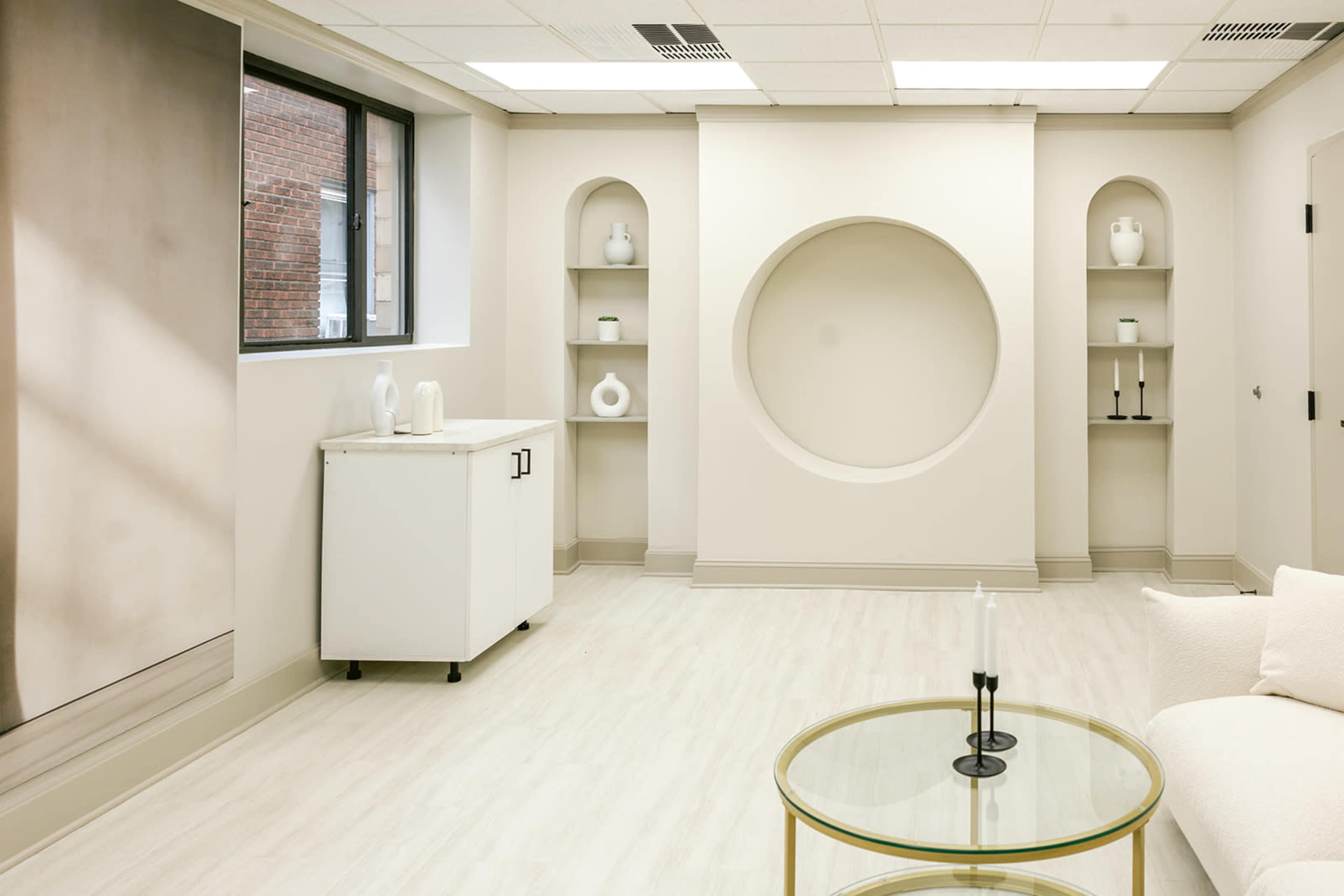 The image shows a minimalist interior room with light-colored walls, a circular wall feature, built-in shelves, and a small white cabinet.