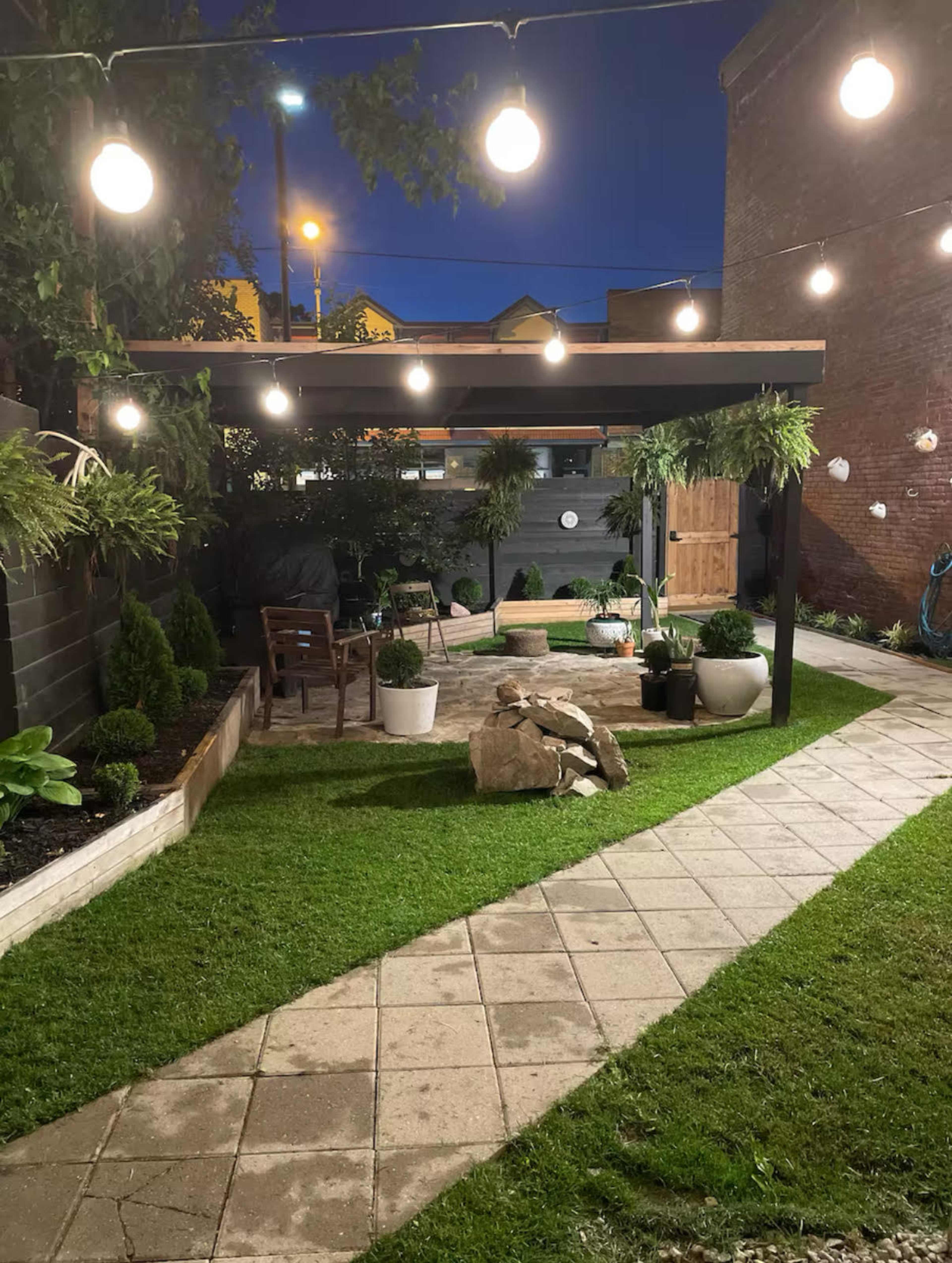 The image shows a well-lit backyard garden featuring a stone pathway, a seating area under a pergola adorned with hanging lights, planters, and lush greenery.