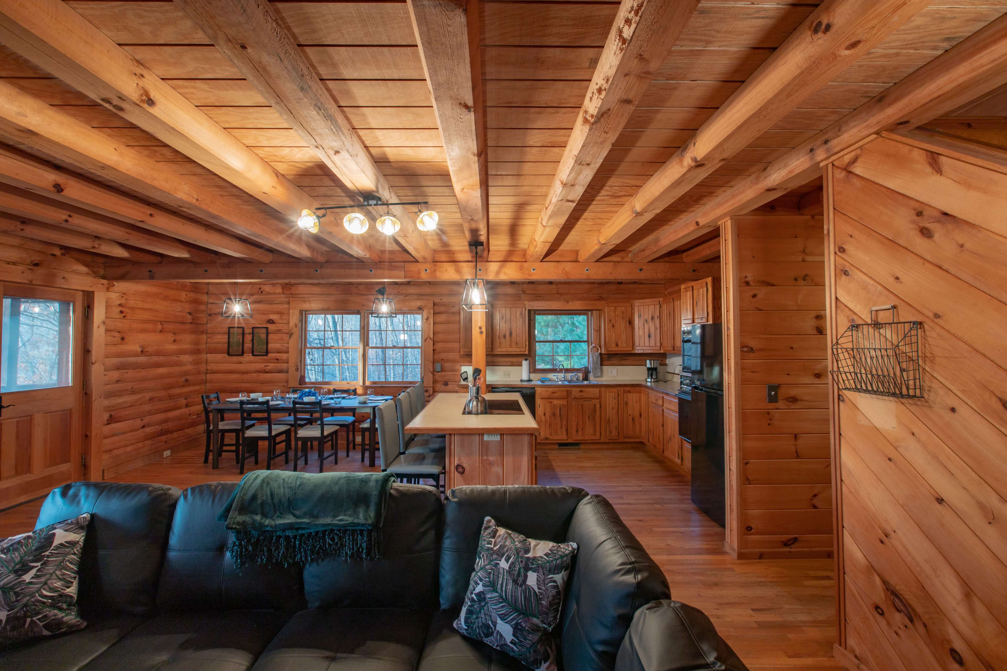 The interior of a wooden cabin features a cozy living area with a black sofa, an adjoining kitchen, and a dining area with a table set for six.