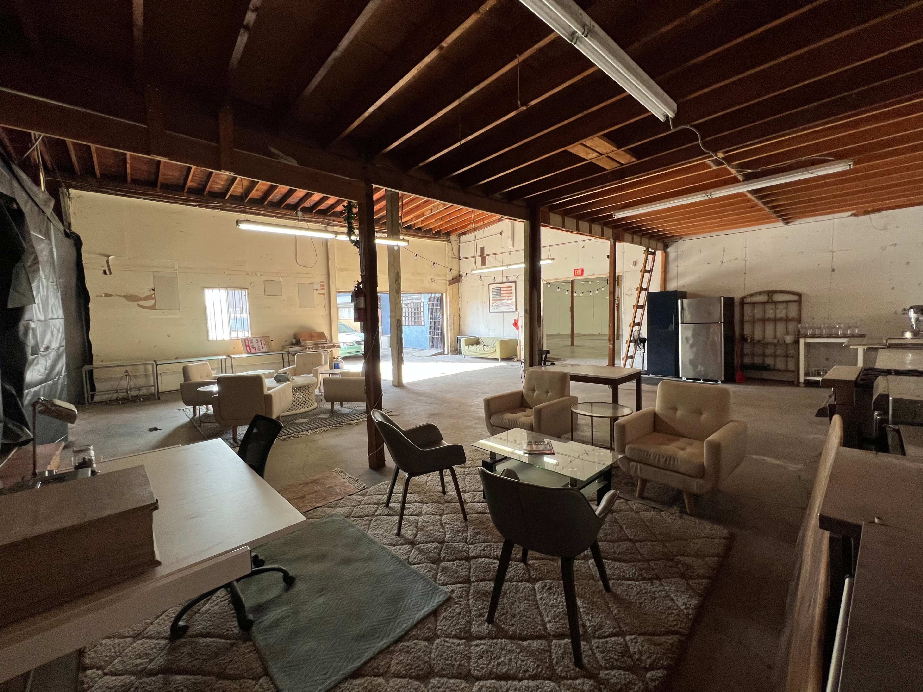 A spacious, unused interior space with wooden beams, a concrete floor, and several tables and chairs arranged throughout.