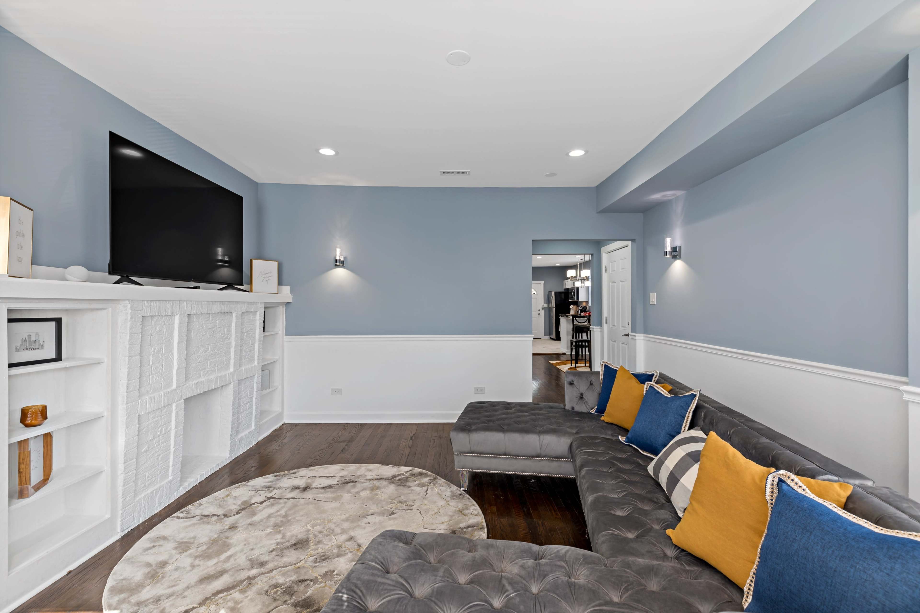 The living room features a sectional sofa with blue and gold cushions, a flat-screen TV on the wall, and a circular rug on a hardwood floor.