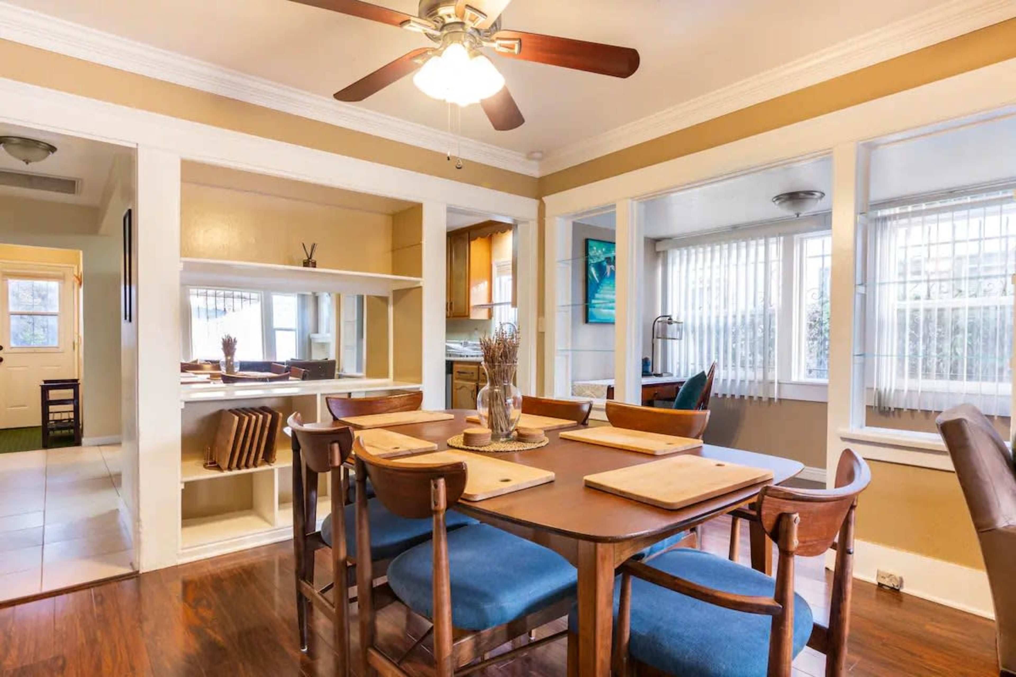 The image shows a dining area with a wooden table set for a meal, surrounded by blue chairs, leading to a kitchen area with large windows and a ceiling fan above.