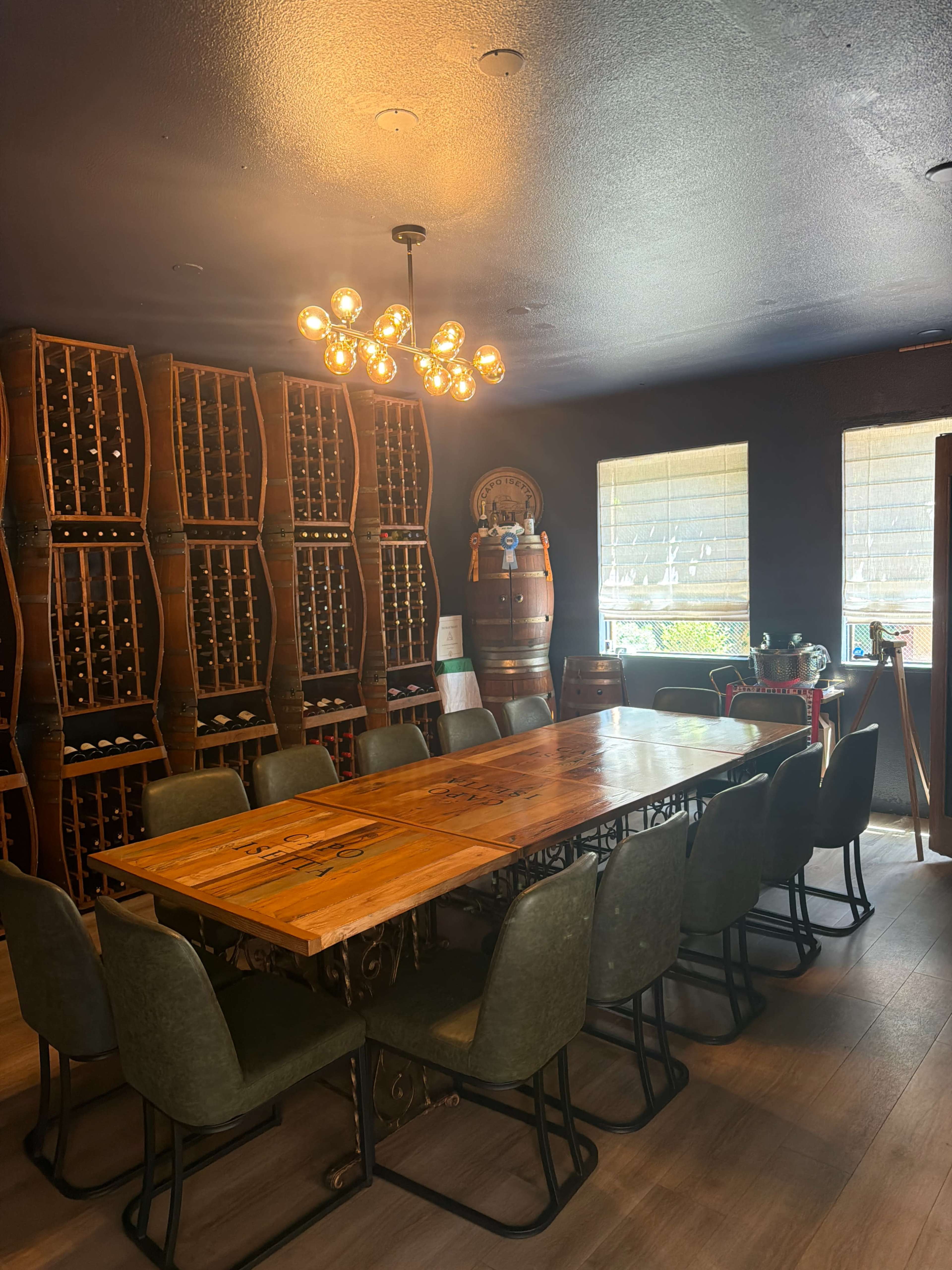 The image shows a spacious dining room featuring a large wooden table surrounded by chairs, with a wall of wine racks and decorative barrels.