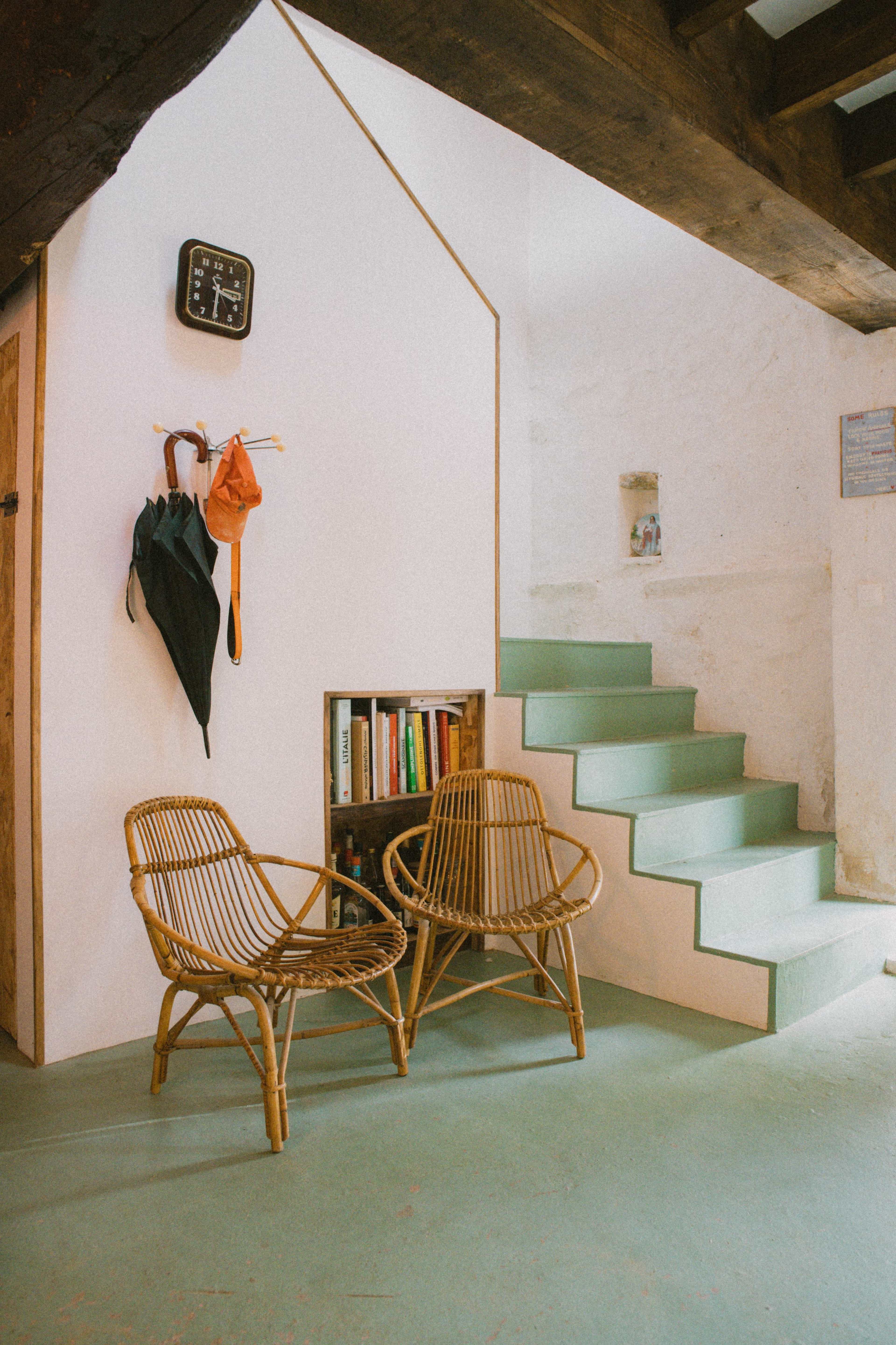 A cozy interior space featuring two wicker chairs beside a staircase, with books displayed in a recessed wooden shelf and a clock on the wall.