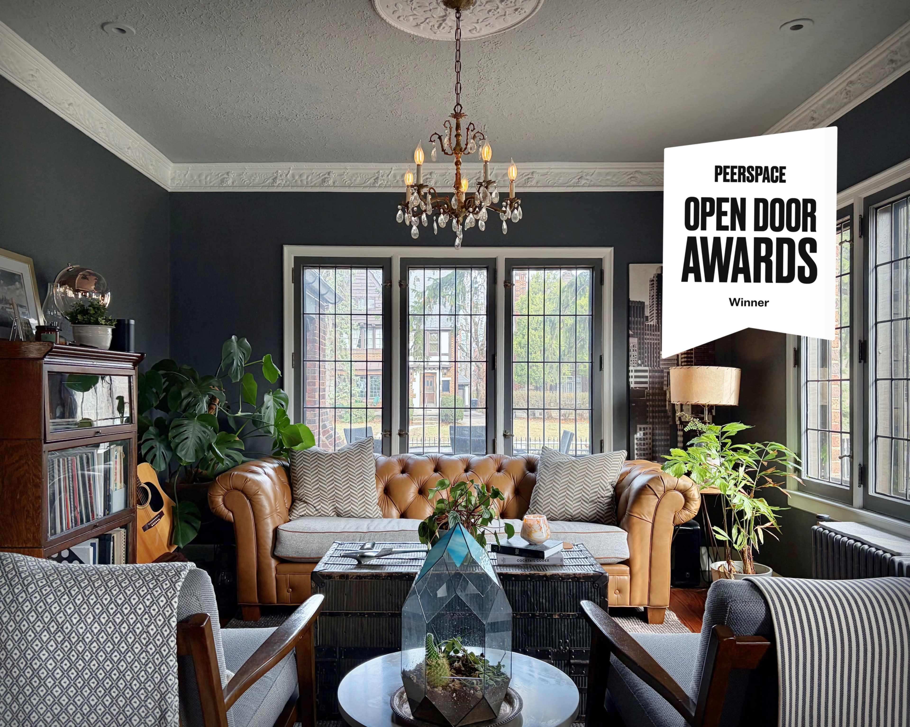 The image shows a cozy living room with a leather chesterfield sofa, a dining table in the center, large windows letting in natural light, and various plants.