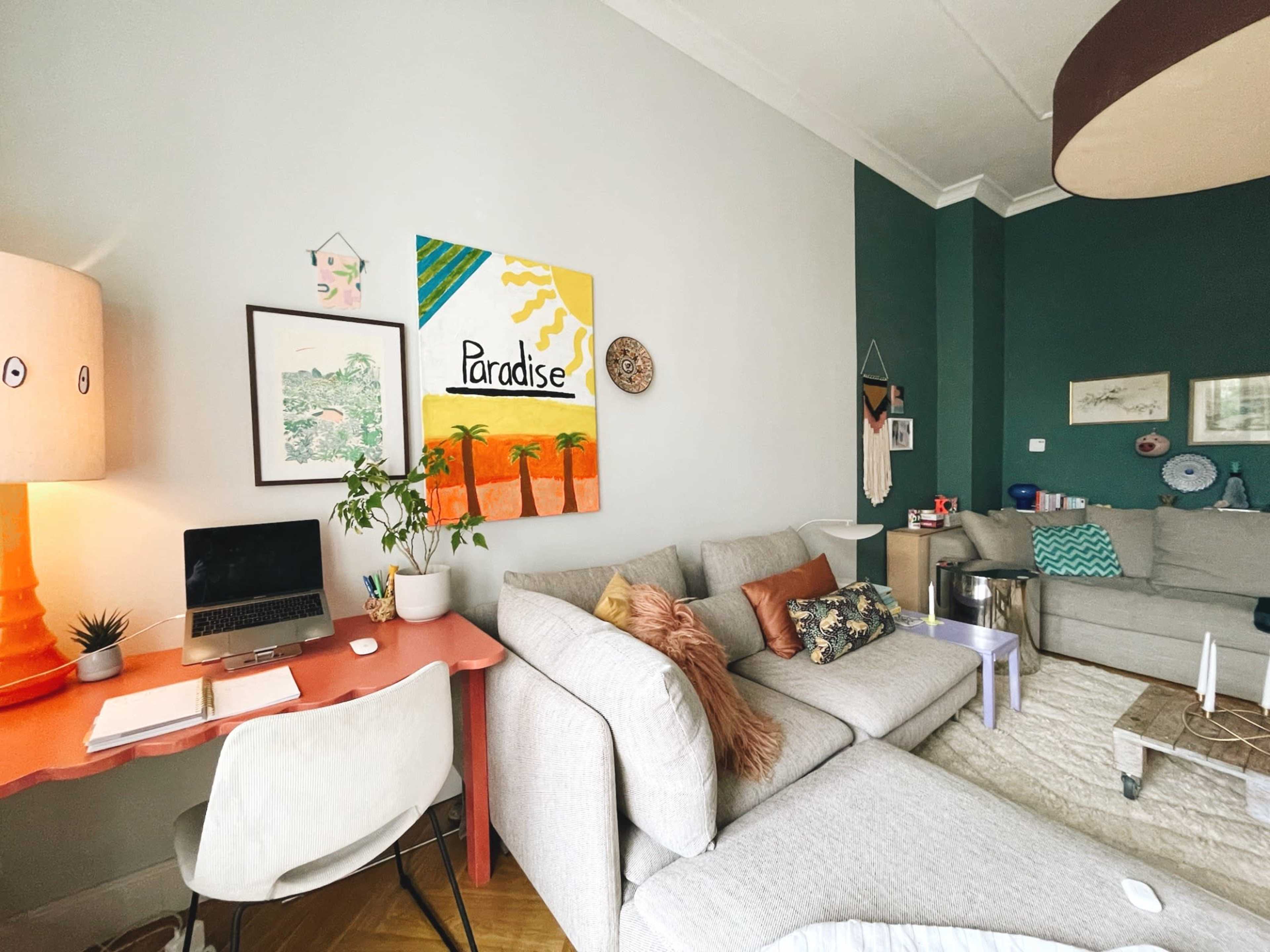 The living room features a light gray sectional sofa, a coral desk with a laptop, and vibrant artwork, including a painting titled "Paradise" on the wall.