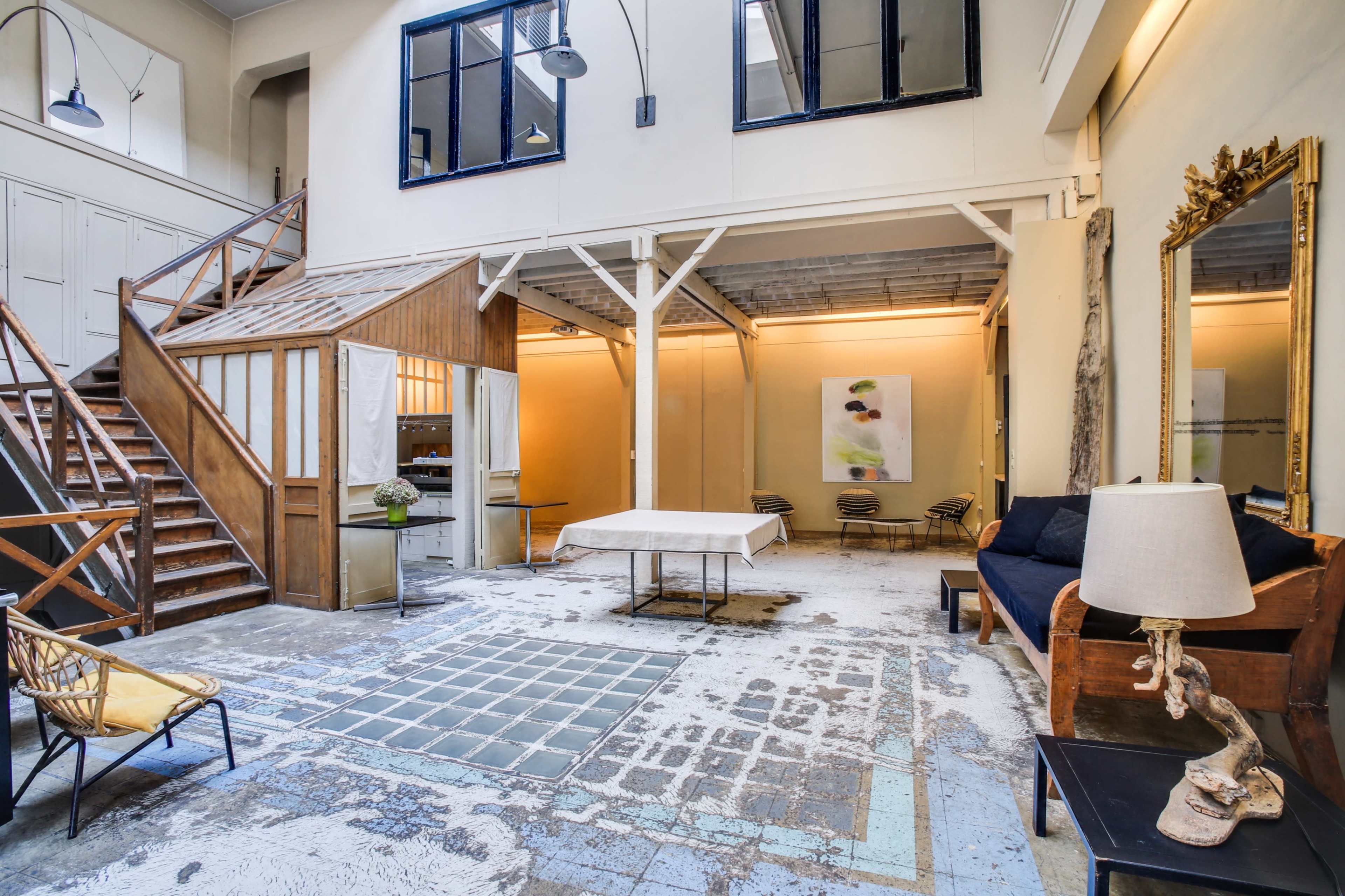 A spacious, industrial-style interior with a staircase, seating area, and large windows allowing natural light to enter.