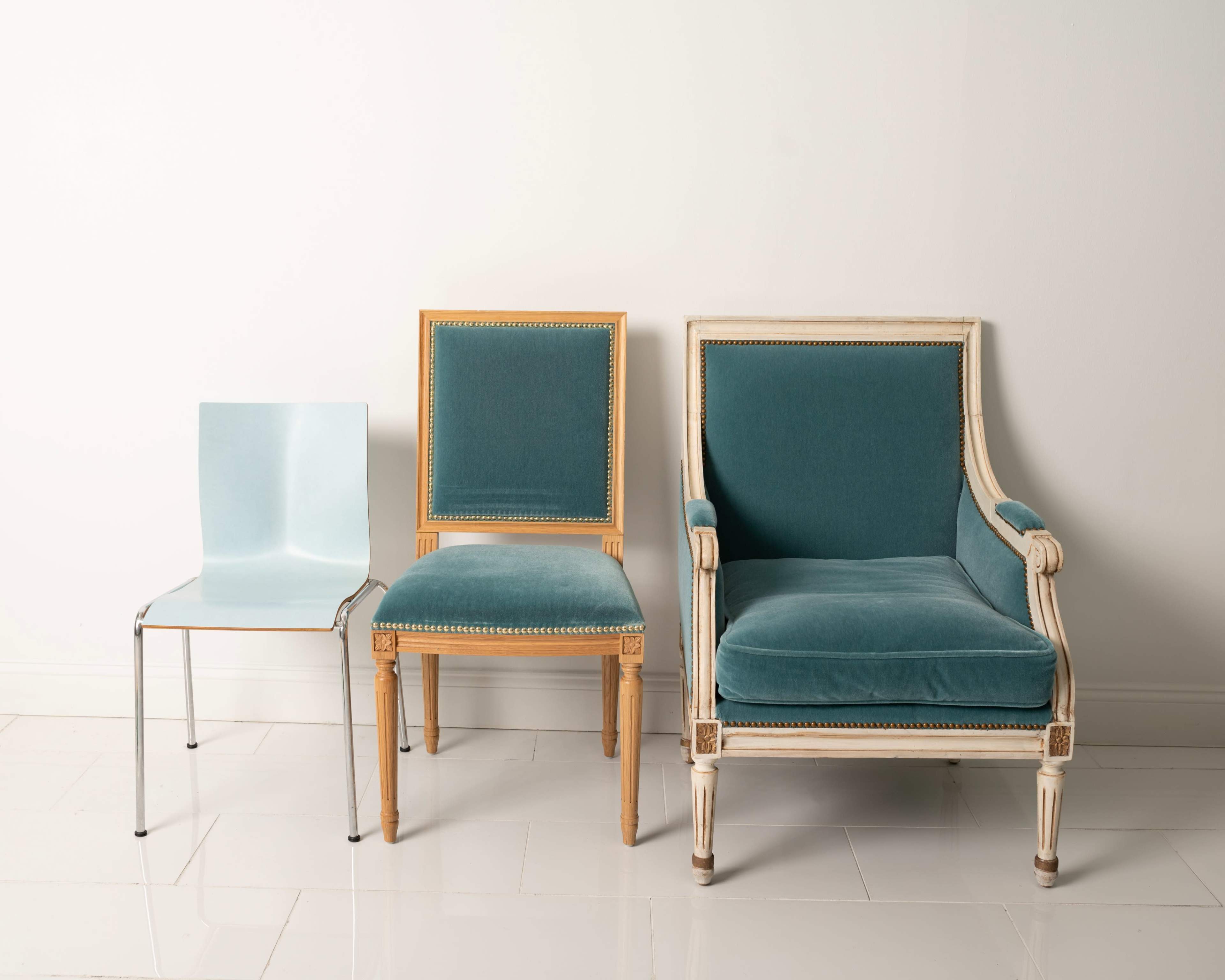 The image features three distinct chairs: a modern clear plastic chair on the left, a wooden chair with a velvet seat in the middle, and a plush upholstered armchair on the right, all positioned against a neutral wall.