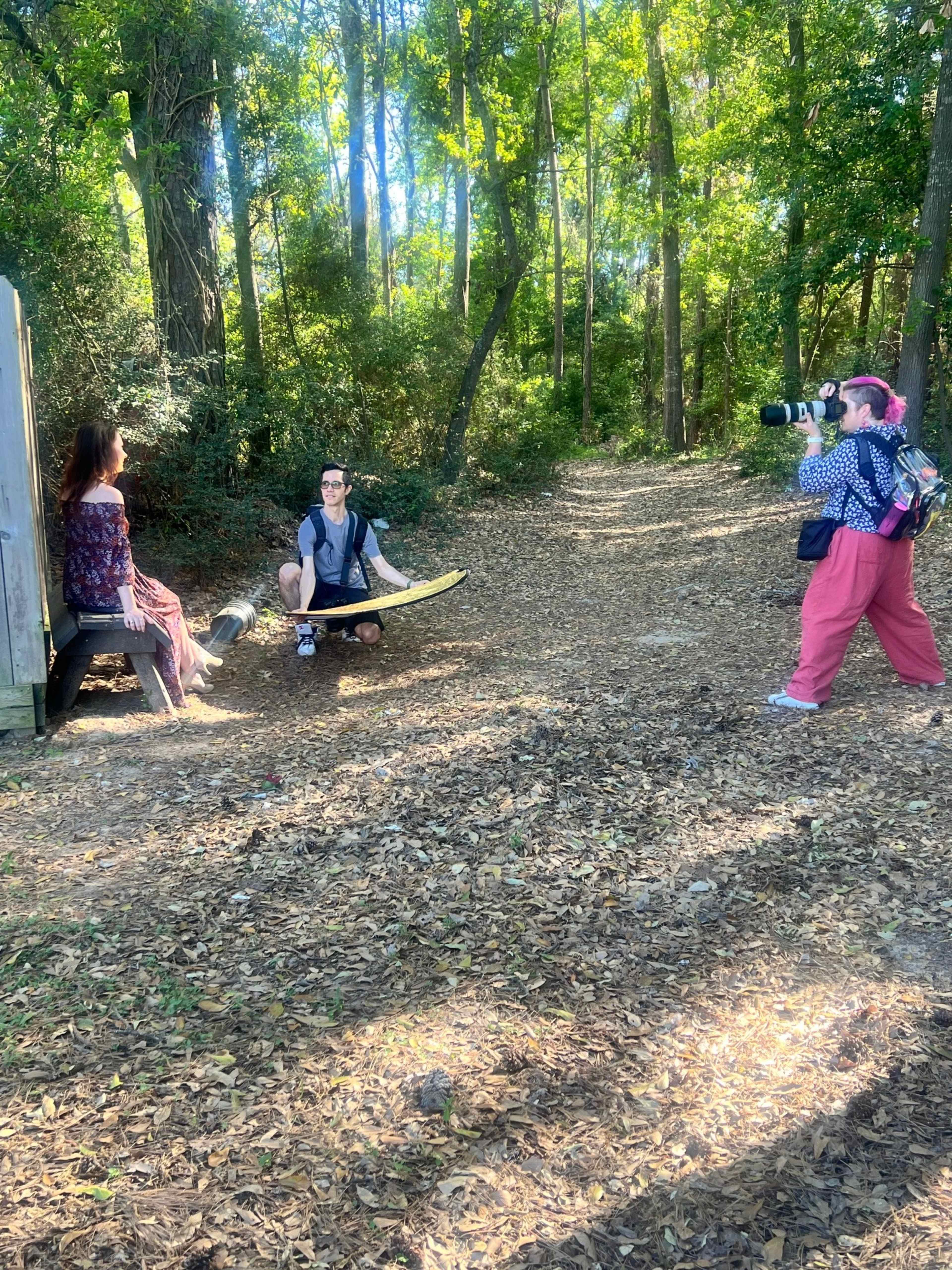 A photographer captures a model sitting on a wooden structure in a wooded area while another person kneels nearby.