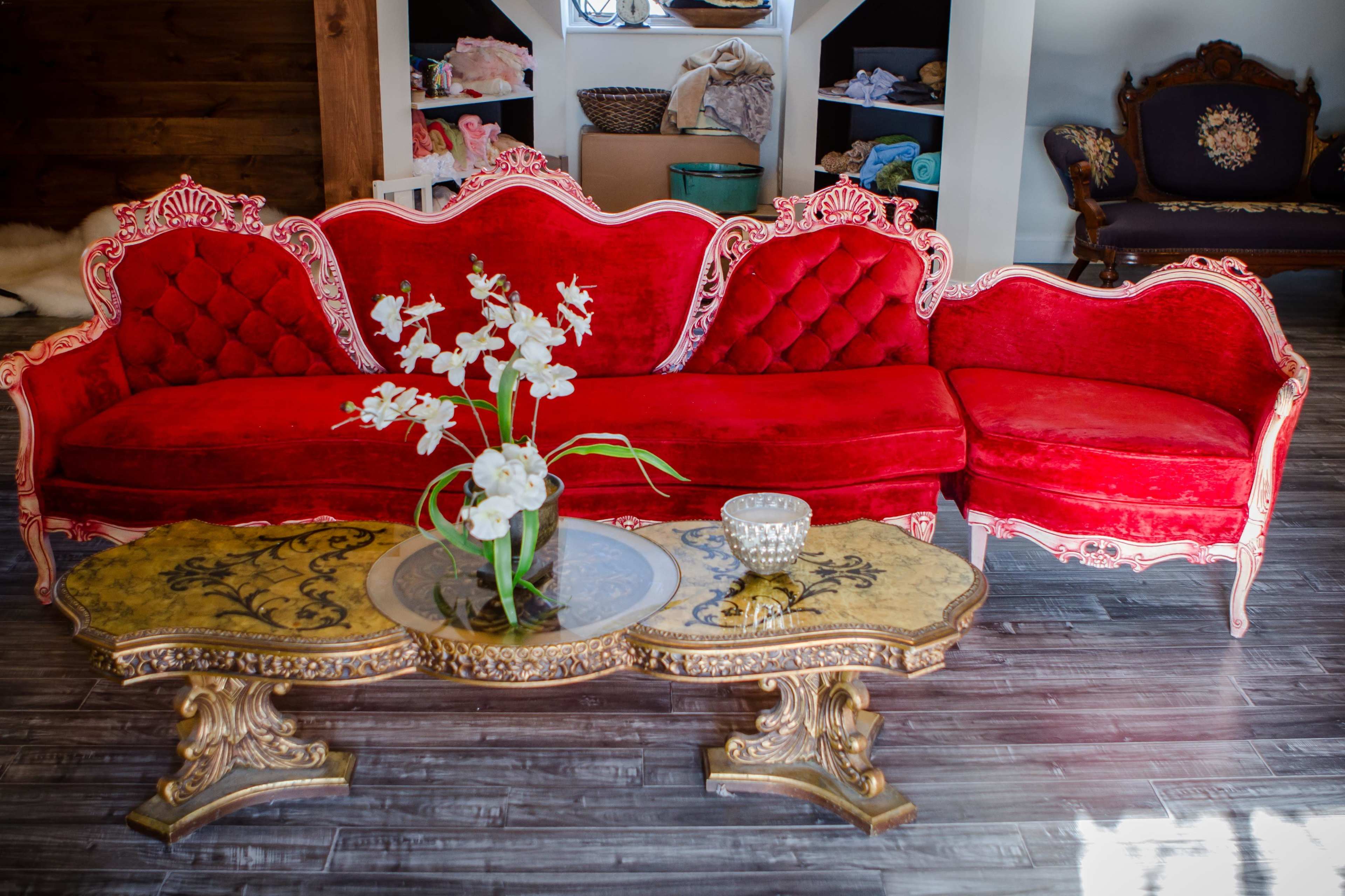 A red velvet couch with ornate wooden trim sits beside a gold-accented coffee table, featuring a floral arrangement.