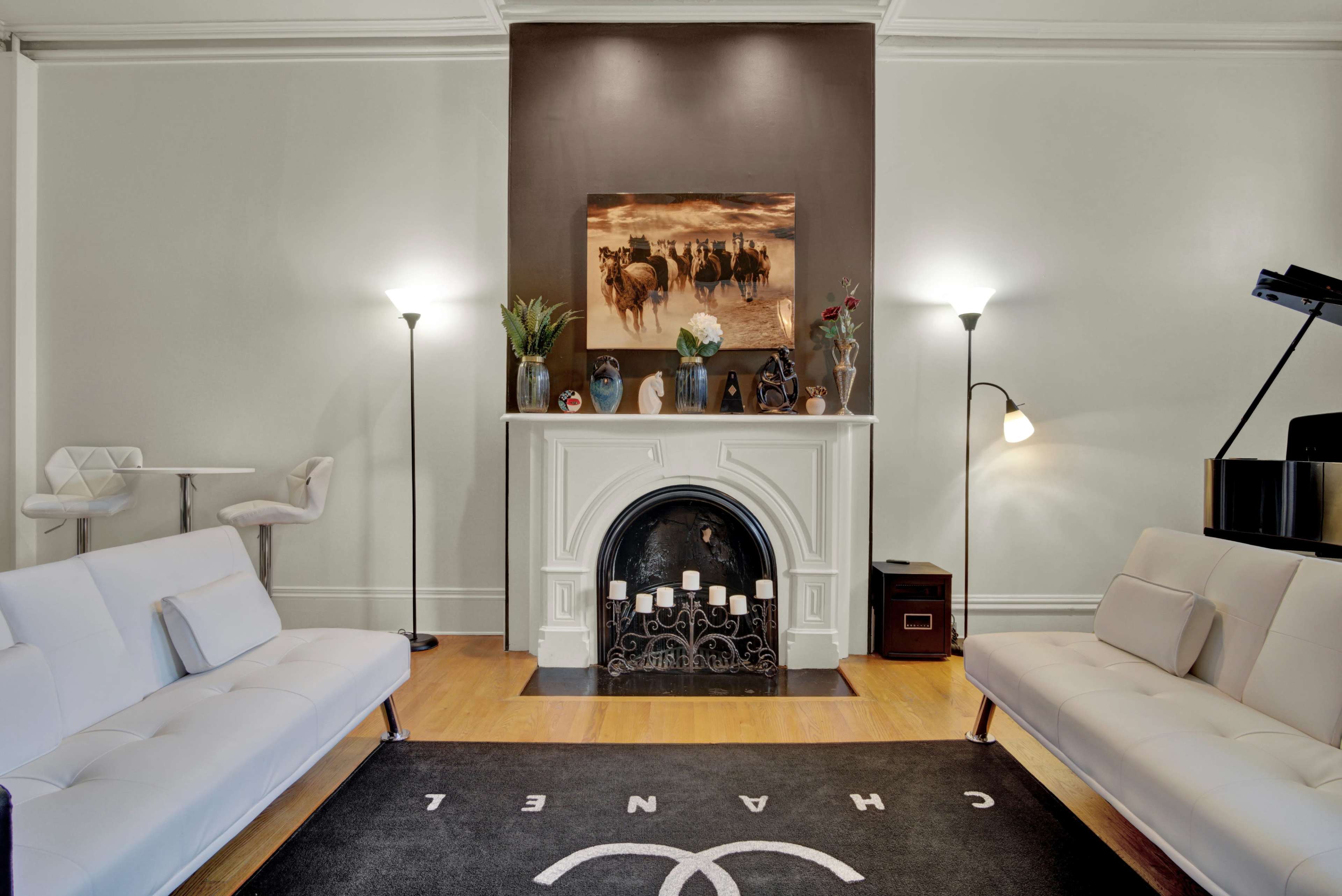 A modern living room features a white fireplace with a painting above it, flanked by floor lamps and two white sofas on either side of a black carpet with a logo.