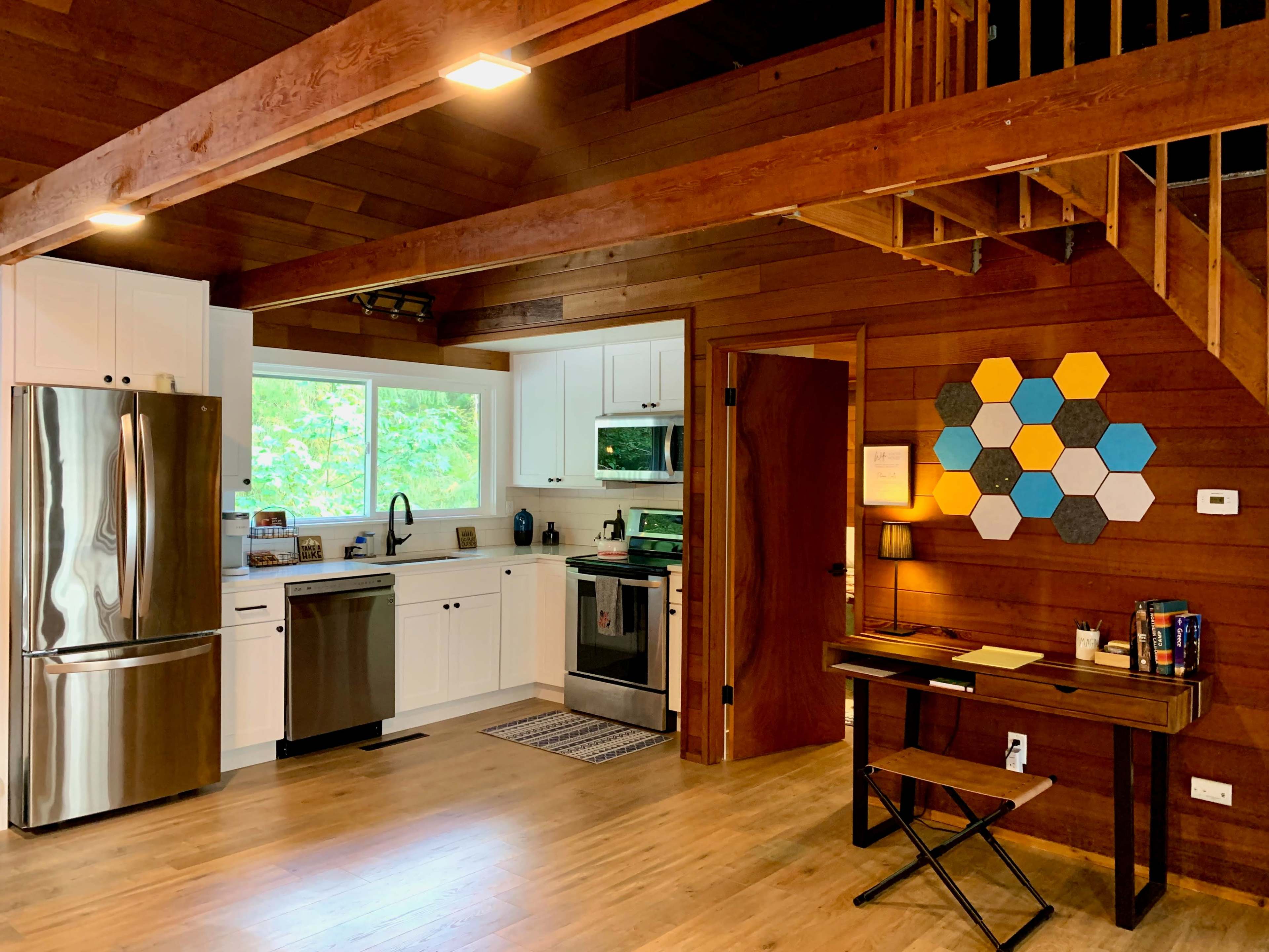 A modern kitchen and workspace area in a wooden interior, featuring stainless steel appliances, a desk, and a staircase.