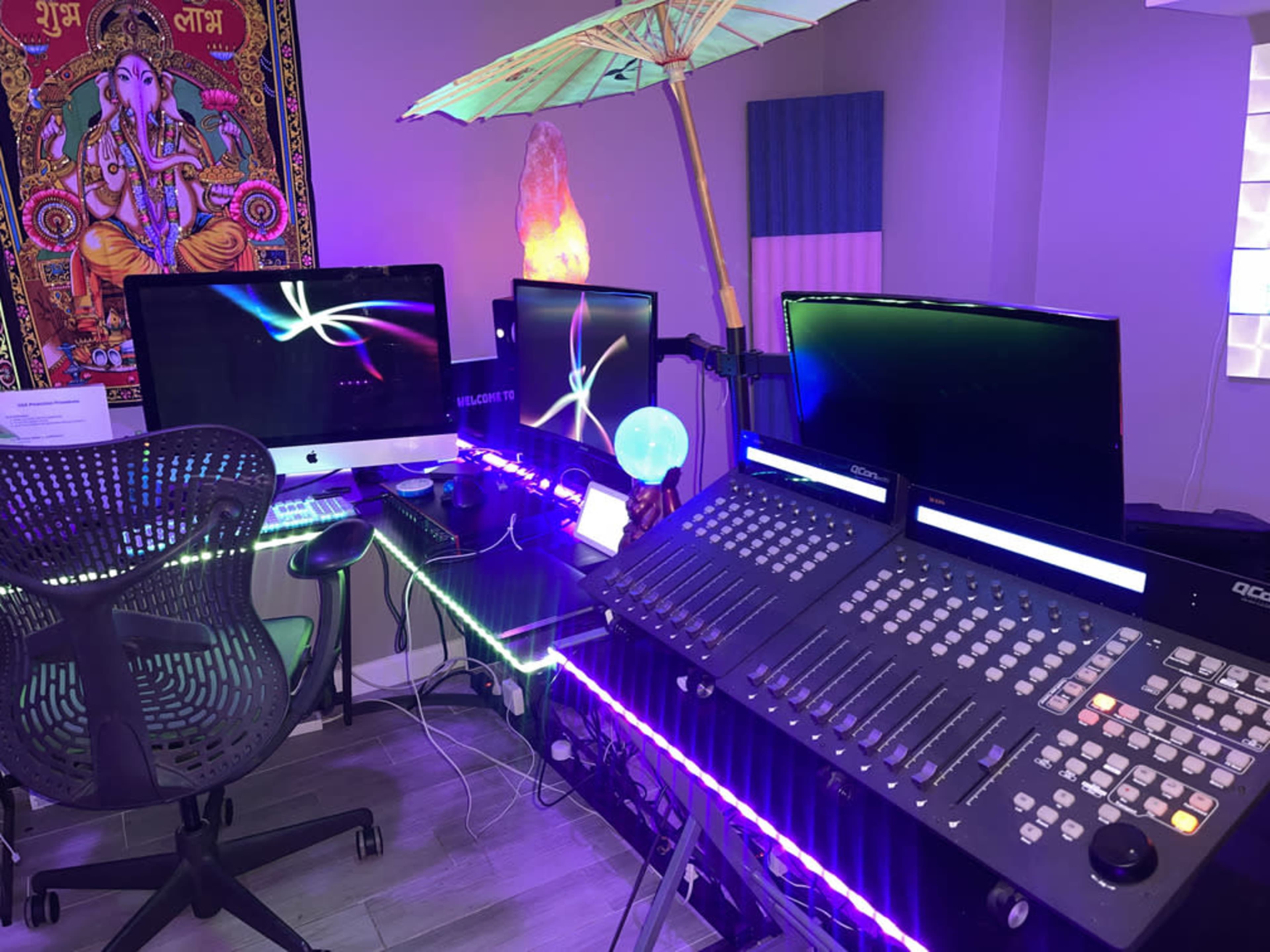 A modern workspace featuring multiple computer monitors, a mixing console, and decorative elements like a parasol and a glowing orb.