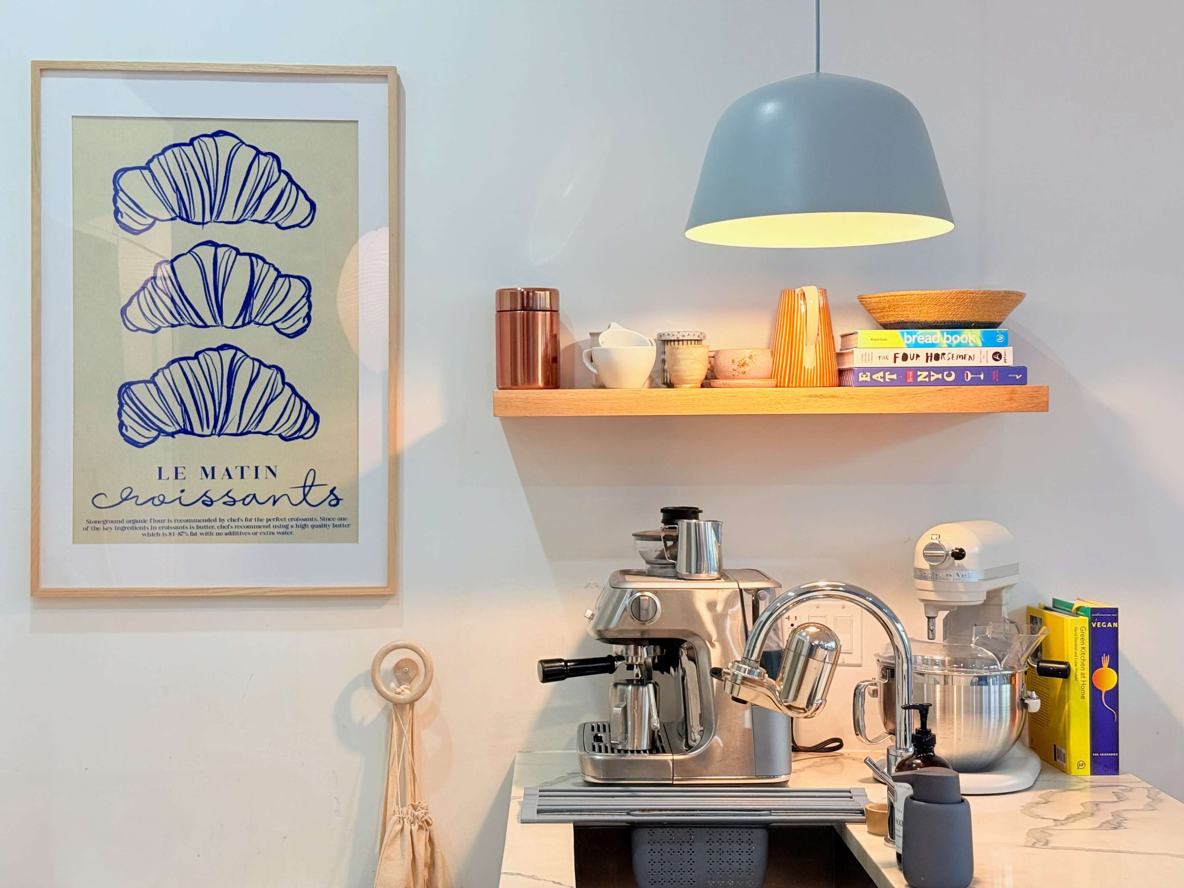 The image shows a modern kitchen corner with a coffee machine, a stand mixer, and a shelf displaying books and decorative items, all beneath a pendant light.
