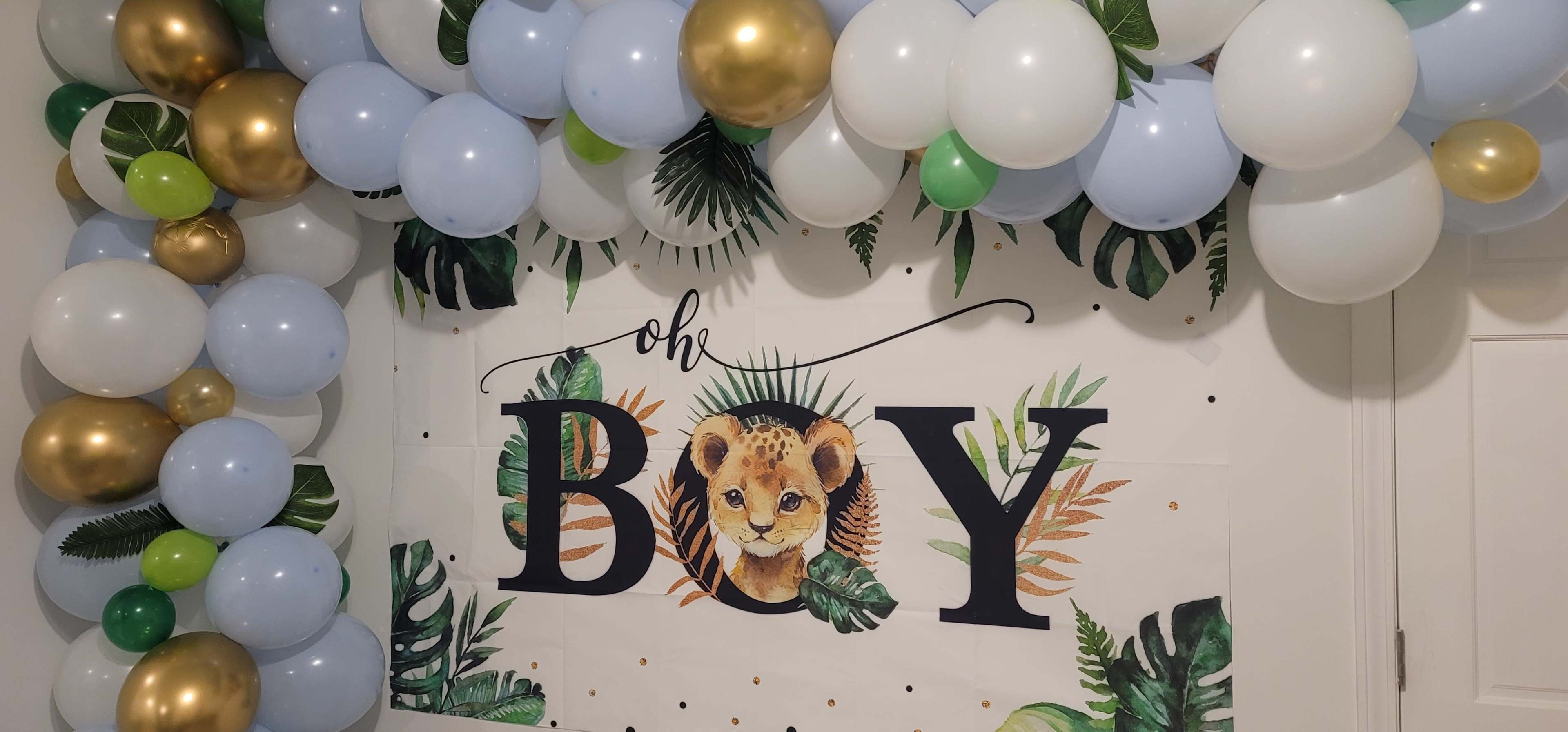A decorated wall with a banner reading "oh BOY" featuring a lion cub and surrounded by a variety of balloons in green, blue, white, and gold.
