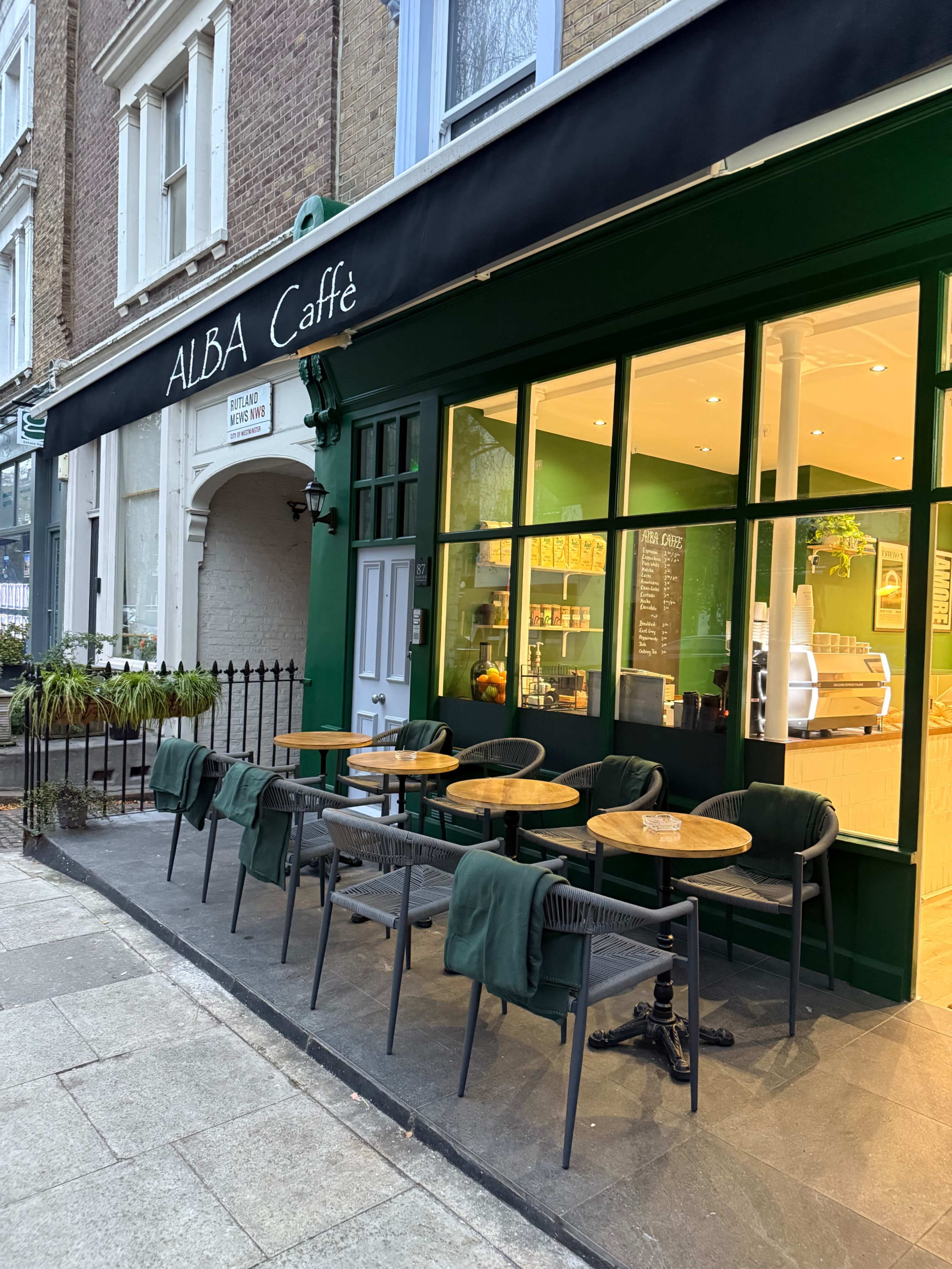 The image shows an outdoor seating area of a café with green walls and wooden tables and chairs in front of a storefront.