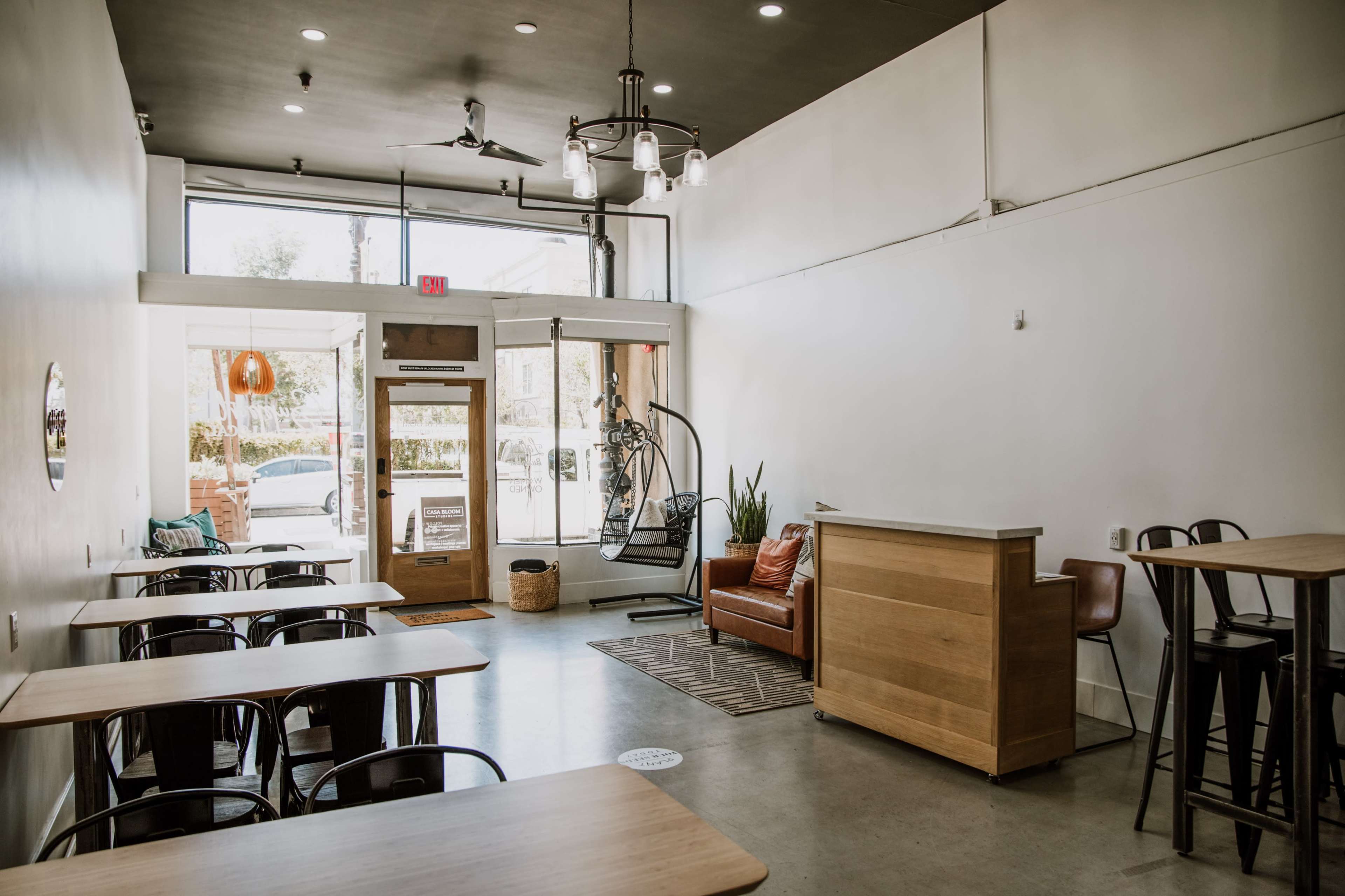 A modern cafe interior with a series of wooden tables, a seating area featuring a leather couch, and large windows letting in natural light.