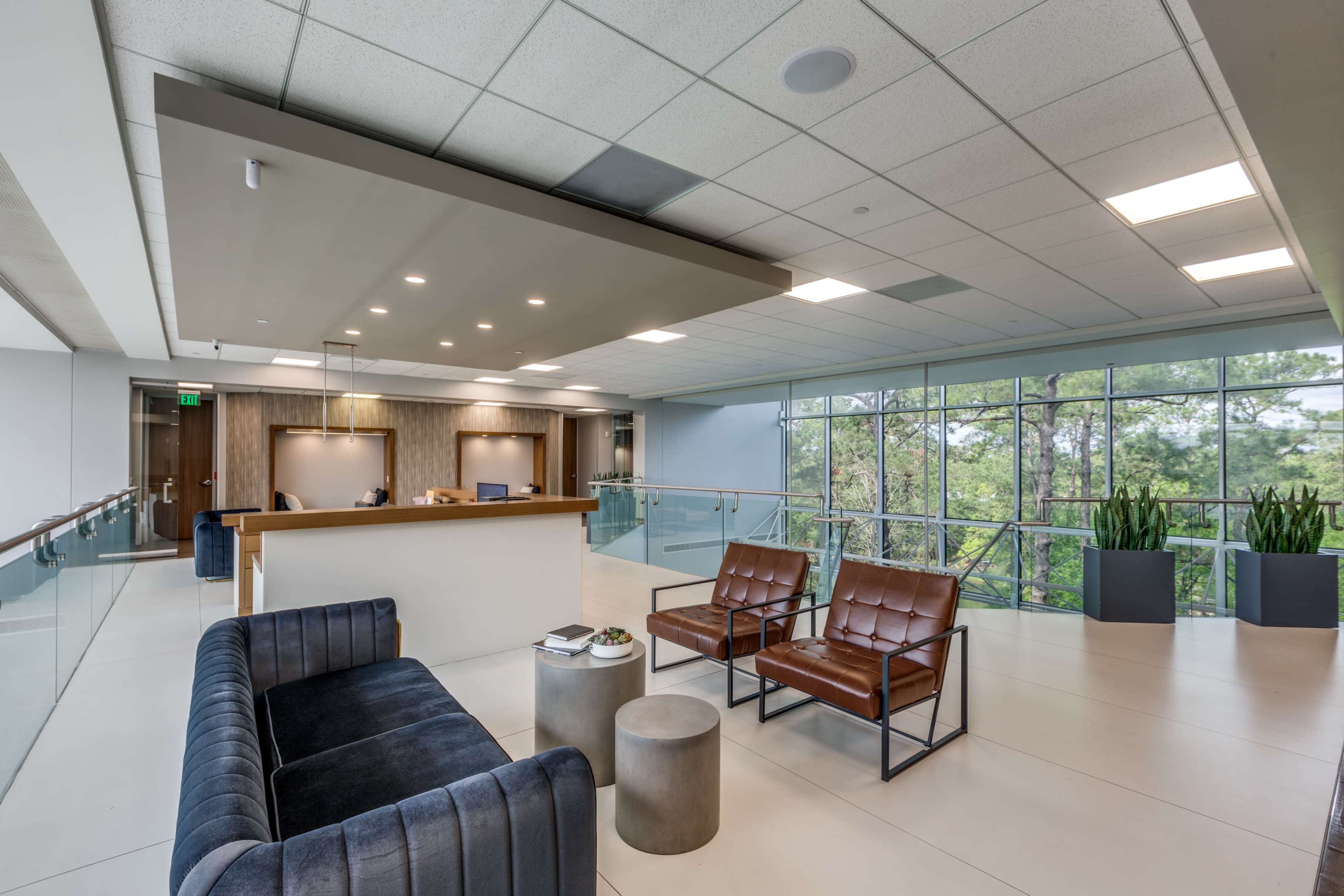 The image shows a modern office lobby featuring a reception desk, two brown leather chairs, a gray side table, and large windows overlooking greenery.