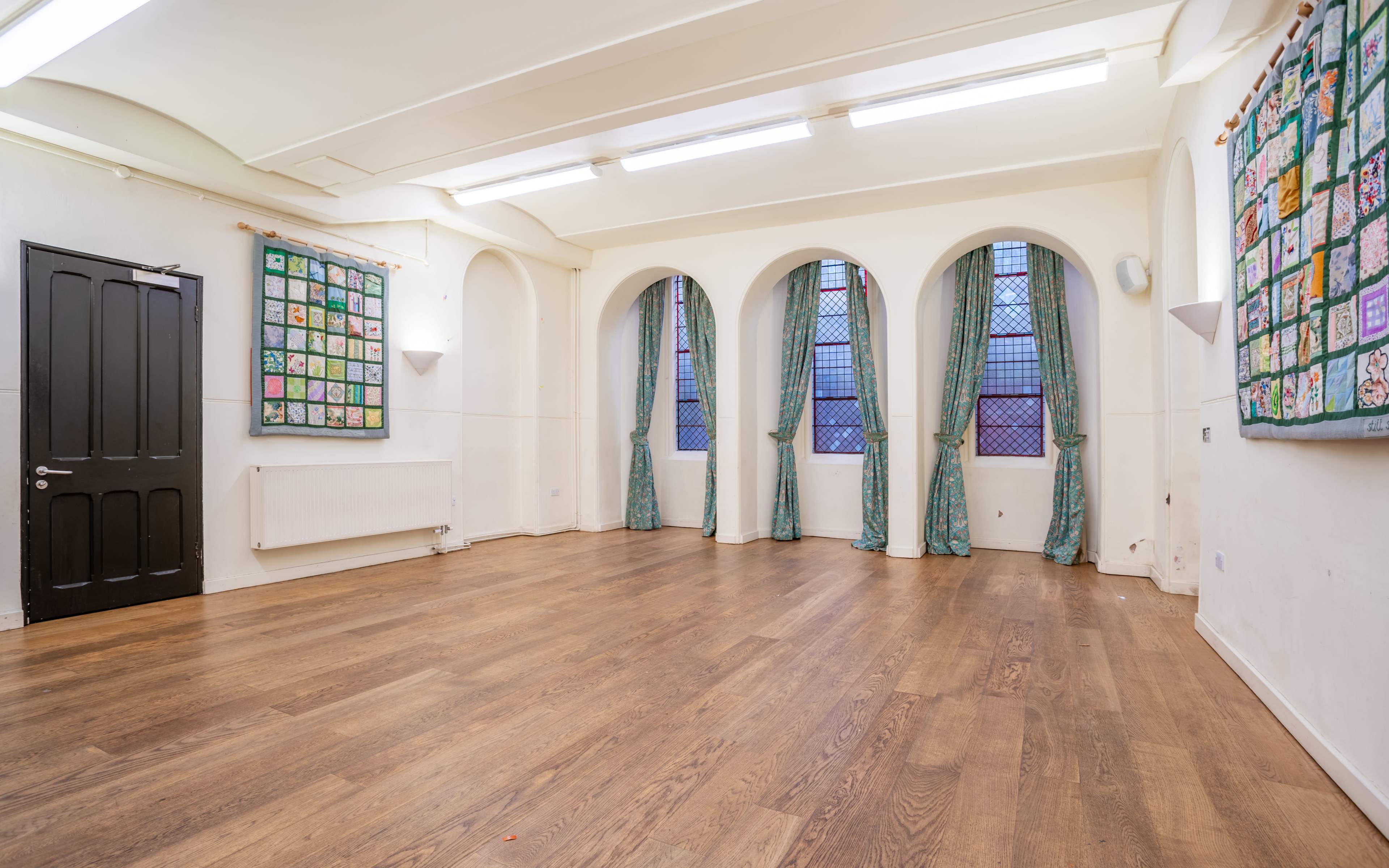 A spacious, empty room with wooden flooring, white walls, and large arched windows draped with green curtains, alongside a wall adorned with a colorful display of framed artwork.