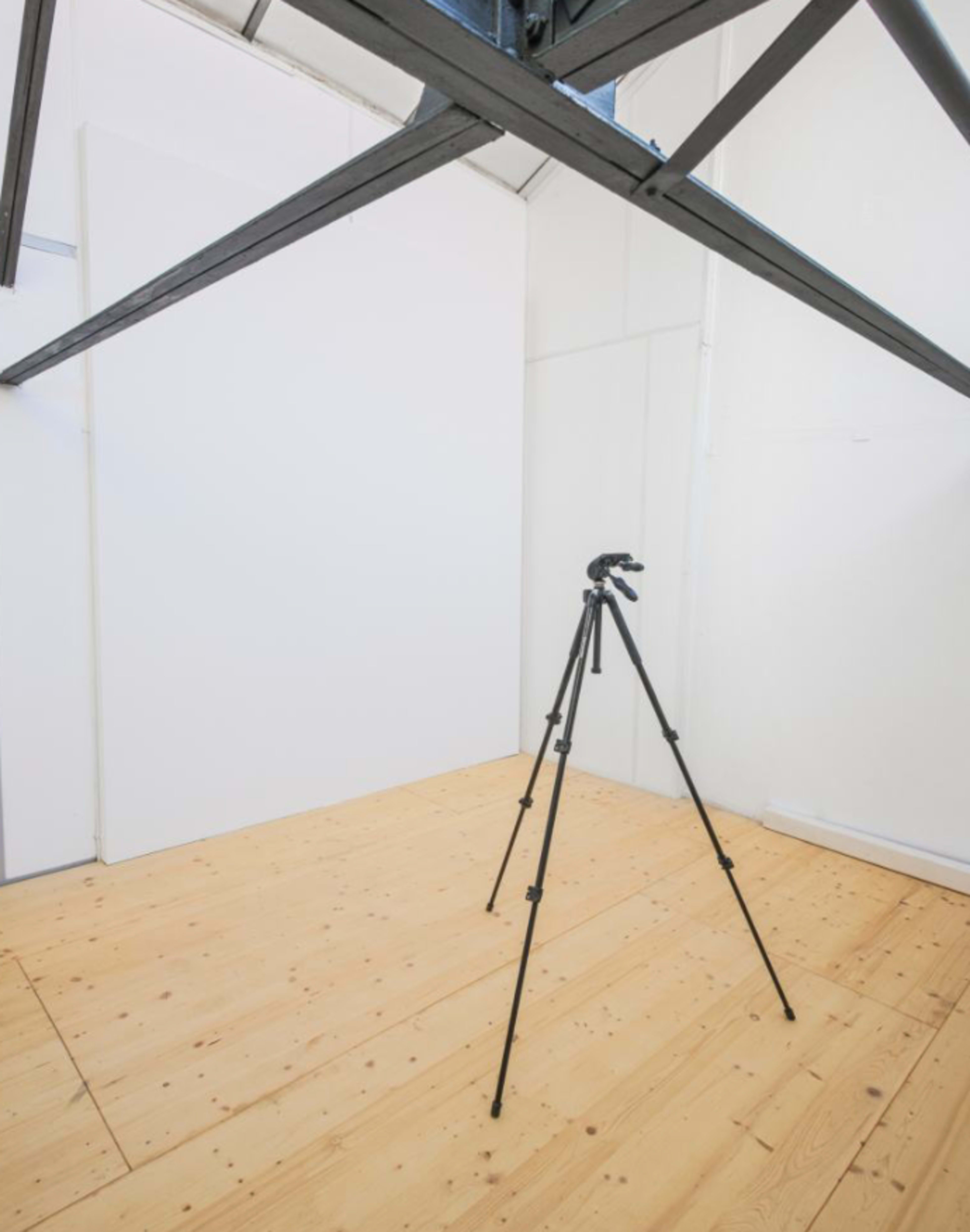 The image shows an empty room with a wooden floor and a tripod positioned in the center, beneath a metal support beam.