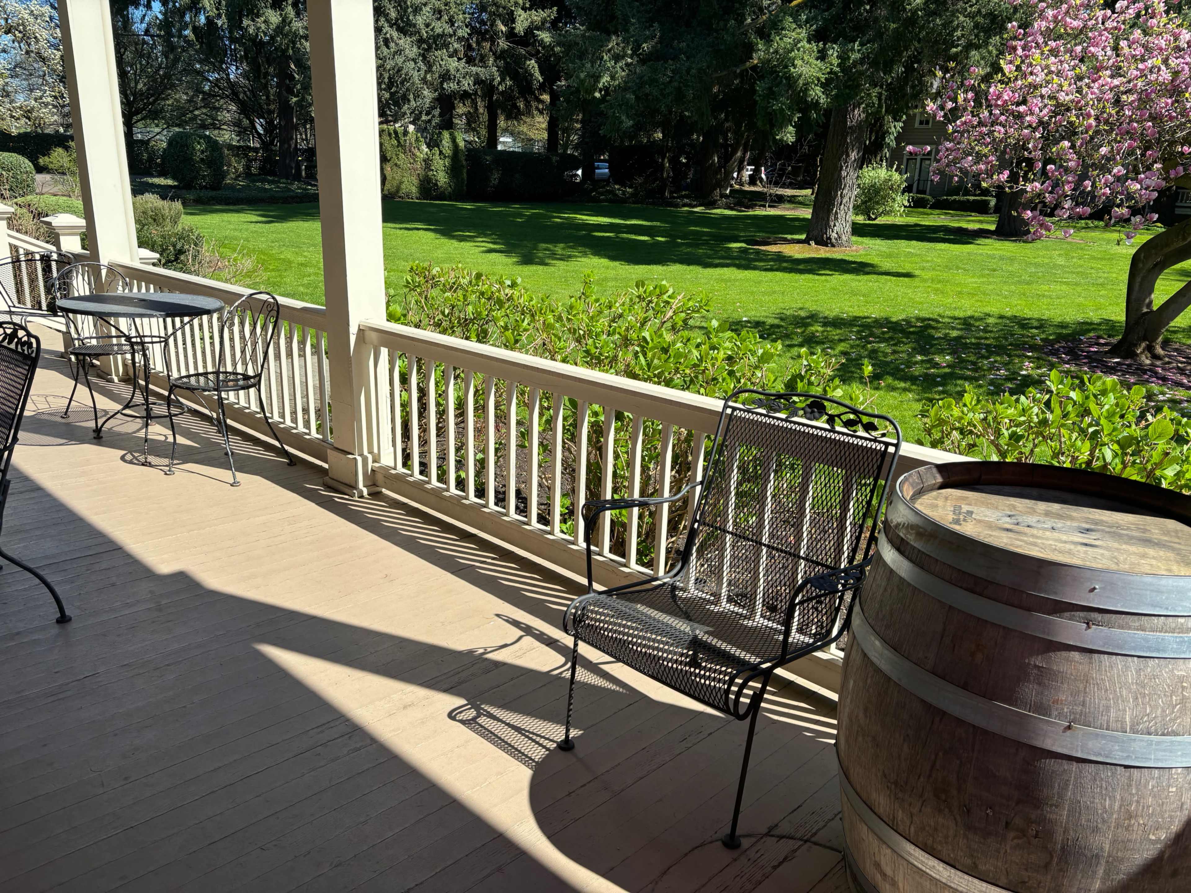A shaded porch features black metal chairs and a wooden barrel beside a green lawn and flowering trees.