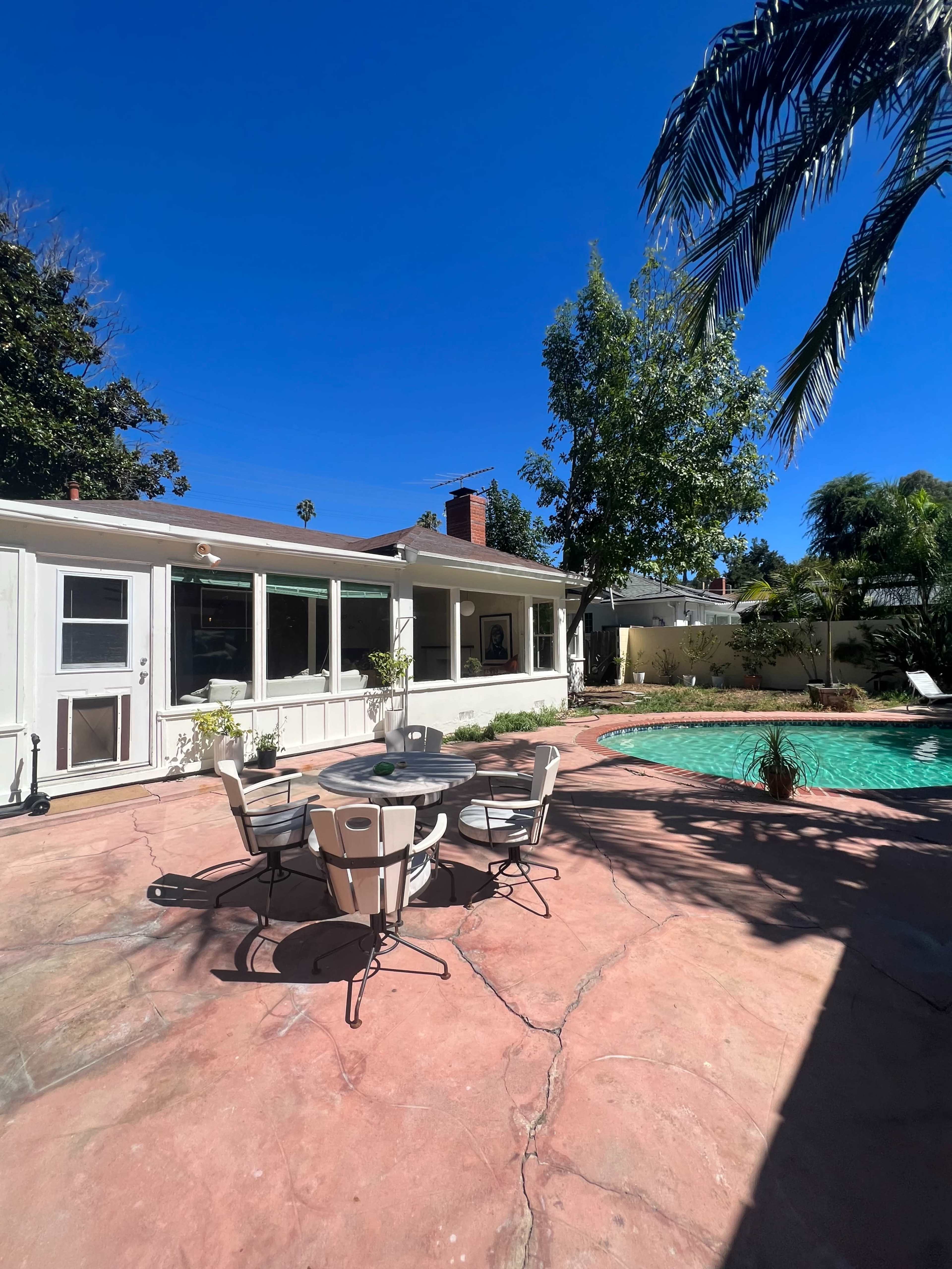 A patio with a round table and chairs overlooks a swimming pool surrounded by greenery and a house in the background.