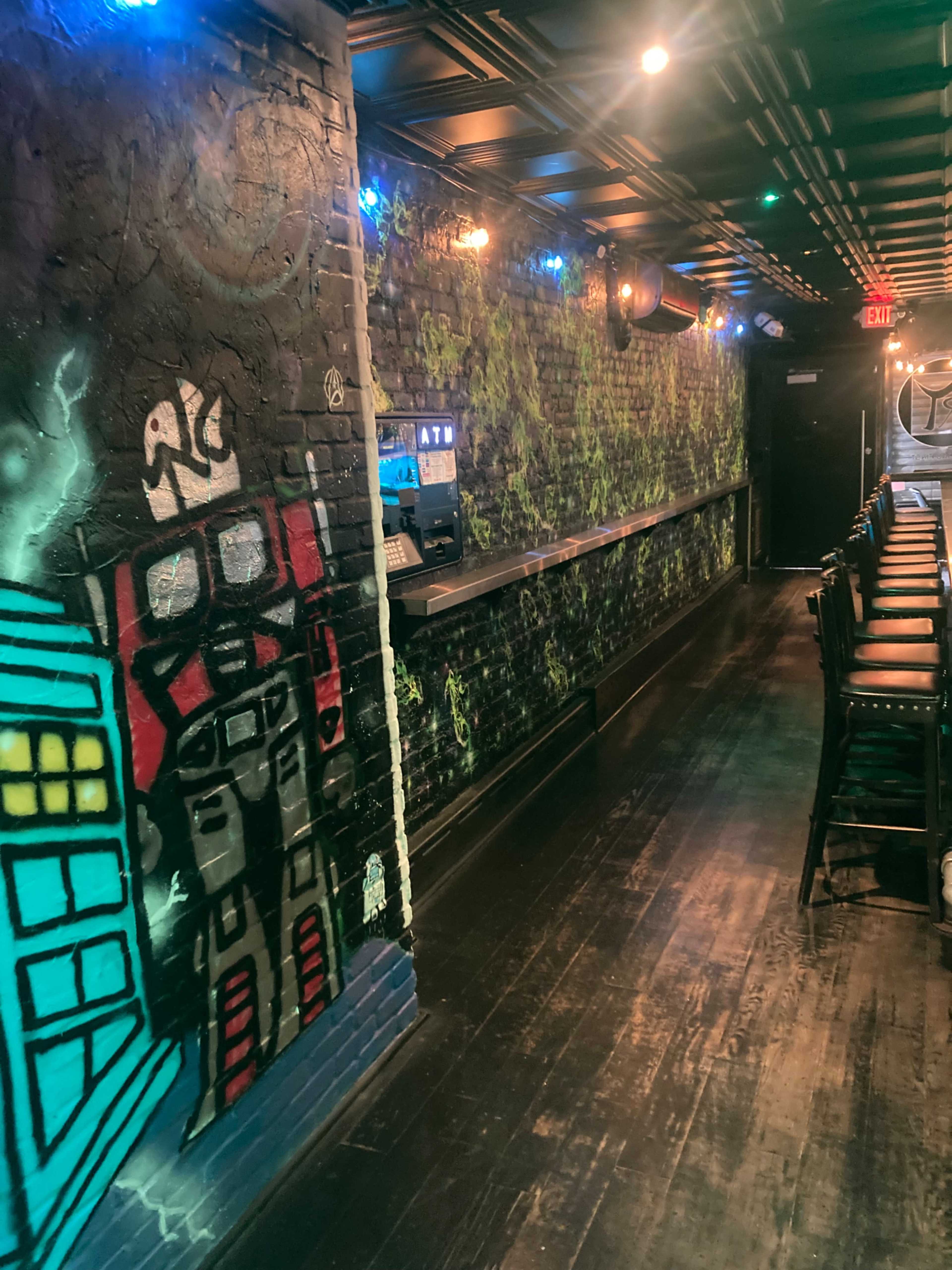 The image shows a dimly lit corridor of a bar, featuring a painted mural of a robot and colorful LED lights along the ceiling.