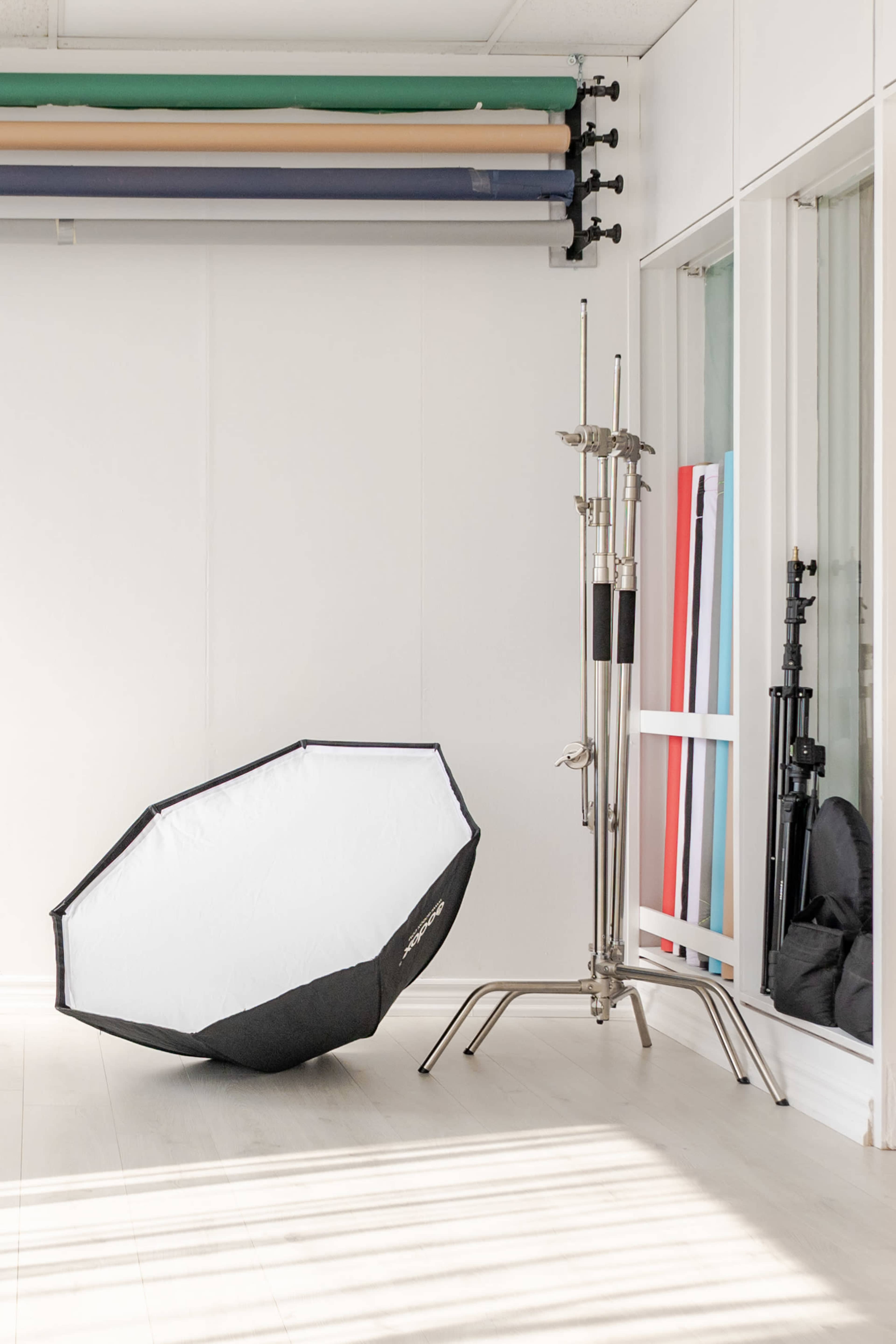 The image shows a photography studio with a large umbrella softbox lying on the floor and several backdrop rolls hanging on the wall in the background.