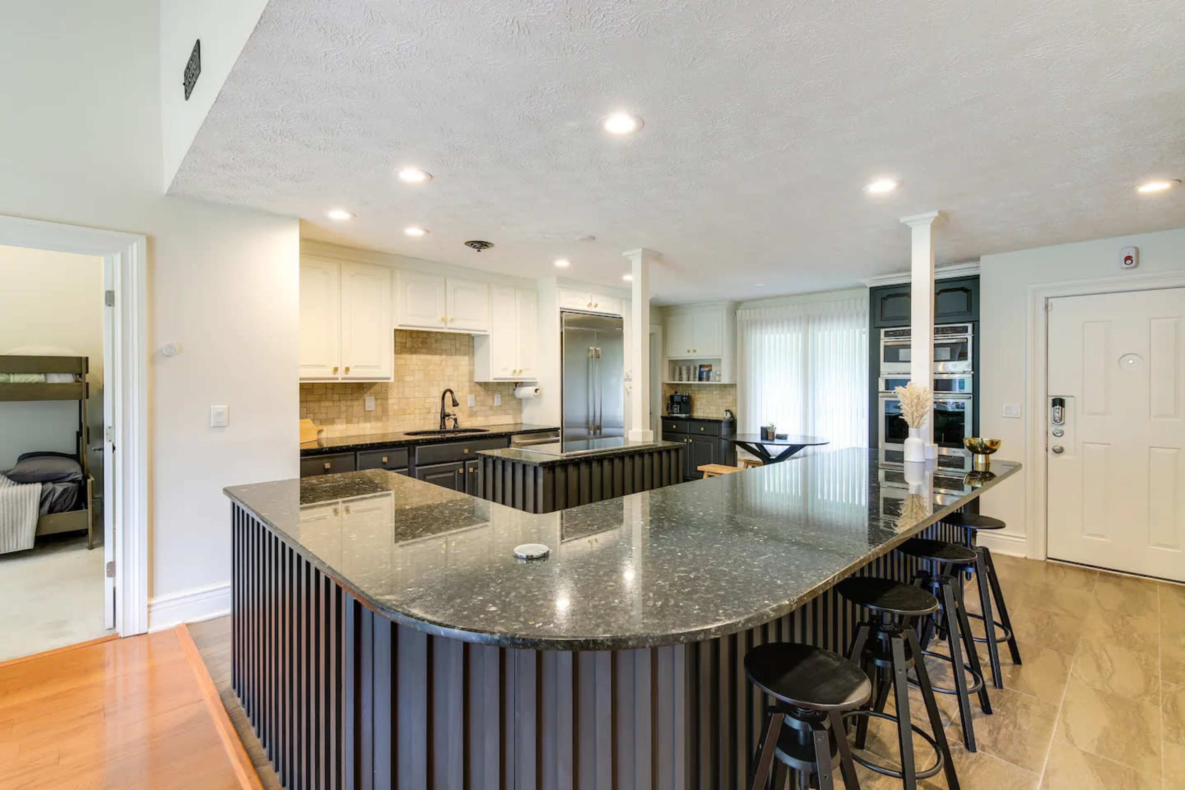 A modern kitchen with a large, dark granite island, black stools, white cabinetry, and a view of a dining space through an open doorway.